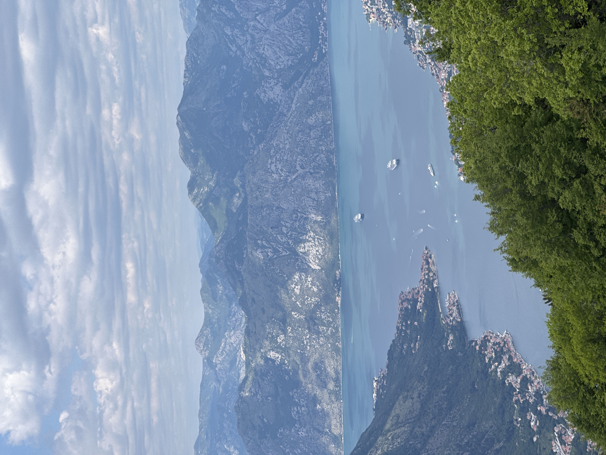 The image depicts a scenic view of Kotor Bay in Škaljari, Montenegro. Mountains rise on the left, gradually descending towards the bay. The bay itself is a deep blue color with several boats visible. The shoreline on the right and in the foreground is dotted with buildings, likely residential. Dense green vegetation is present in the foreground on the right. The sky is light blue with striated clouds.