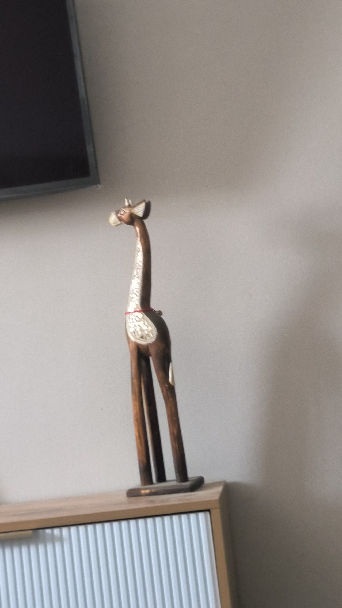 A wooden decorative giraffe figurine stands on top of a light-colored wooden cabinet with white ribbed drawer fronts. The giraffe is brown with white decorative inlay on its neck and body, and a thin red cord around its neck. It is positioned on the right side of the cabinet, against a plain, light gray wall. A portion of a dark-colored television screen is visible in the upper left corner of the image, mounted on the wall above and to the left of the giraffe. The scene appears to be indoors, likely in a living room or bedroom. The specific location in the city of Kovrov, Russia, cannot be determined from the image alone.