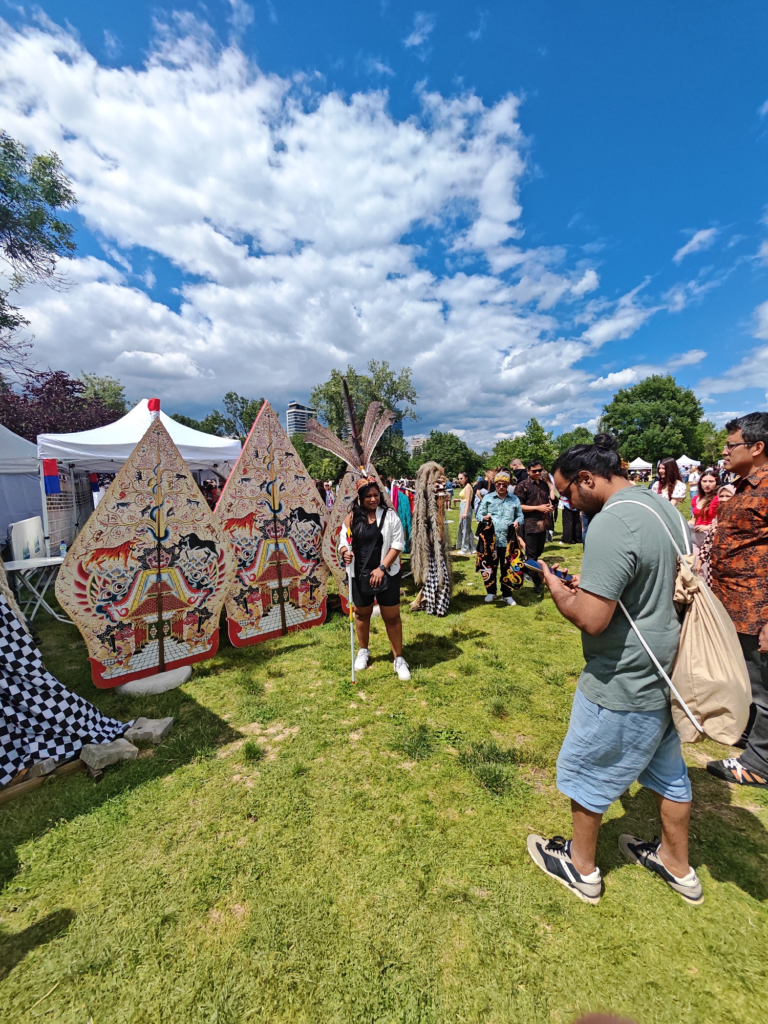 This is a medium-shot, eye-level outdoor photograph taken on a sunny day with a partly cloudy blue sky. The setting appears to be a public event or festival in a park-like area, possibly in the city of Sofia, Bulgaria, given the architecture visible in the background. In the foreground, a man wearing a green t-shirt and blue shorts is looking down at his phone. To his left are two large, intricately decorated puppets, likely Wayang Kulit from Indonesian culture. A woman wearing a black top, black shorts, and a headdress with feathers stands between the puppets. She is holding a long stick. Other people are visible in the midground and background, mingling and observing the displays. Tents, possibly for vendors, are set up behind the puppets. The ground is covered in green grass, and trees and buildings are visible in the distance.