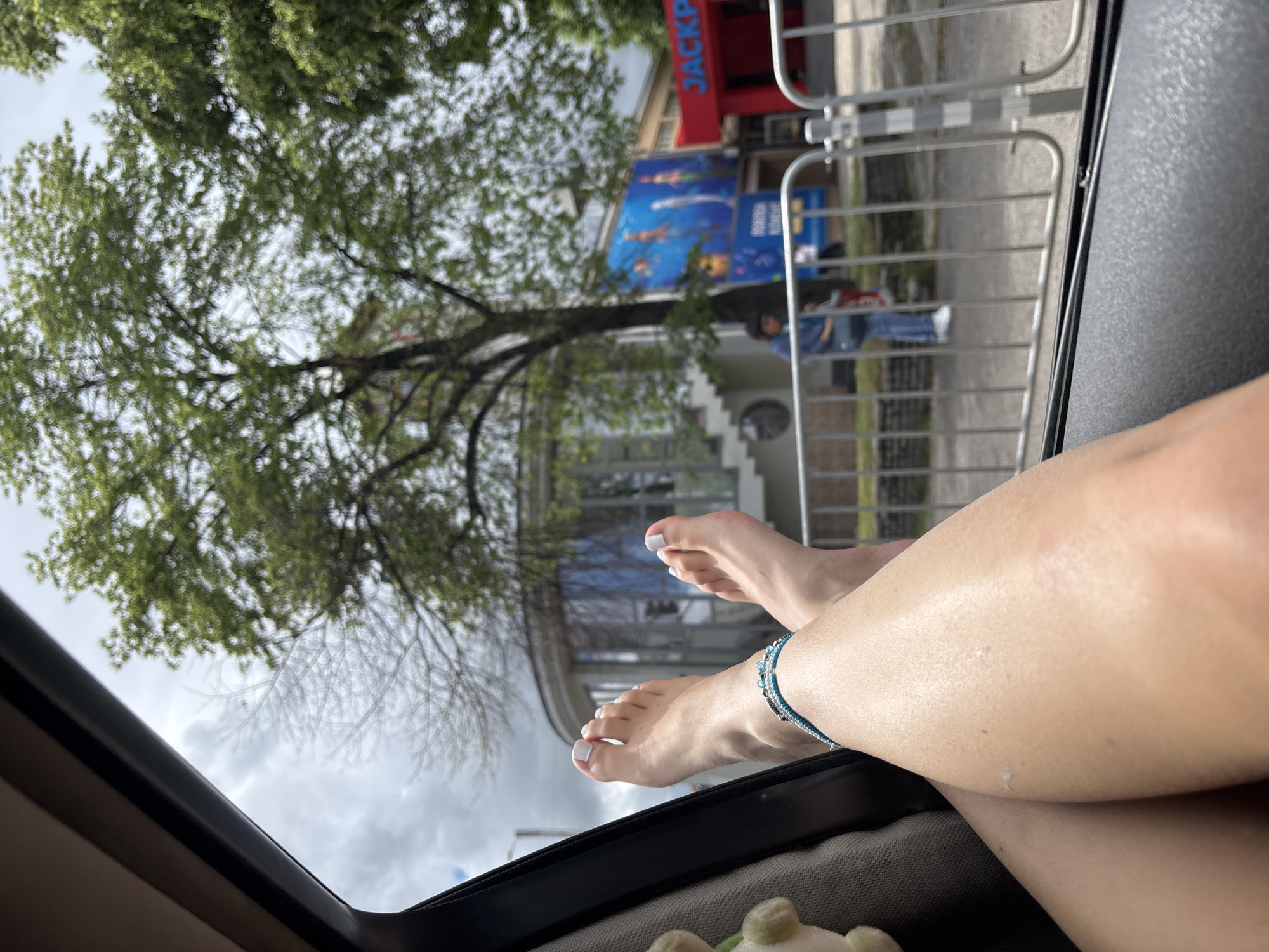 The image, taken from within a car, captures a person's bare feet and lower legs extended casually out of a window or sunroof. The left foot displays light-colored nail polish, and a blue and silver beaded anklet adorns the right ankle. The pose suggests relaxation.
The scene outside the car is an urban environment, viewed under an overcast sky. A large tree with green leaves is prominent on the left side of the frame. To the right, behind metal barriers commonly used for crowd control, several buildings are visible. One building features a red sign with the partial text "JACKP," likely part of "JACKPOT." Below it, a blue sign displays blurred images that appear to depict marine life, possibly indicating an aquarium or aquatic-themed attraction, with text too indistinct to read. A blurred figure of a person is also visible behind the metal barriers.
The setting appears to be a city street in Sofia, Bulgaria, during daytime on a cloudy day, emphasizing a moment of leisure amidst urban activity.
