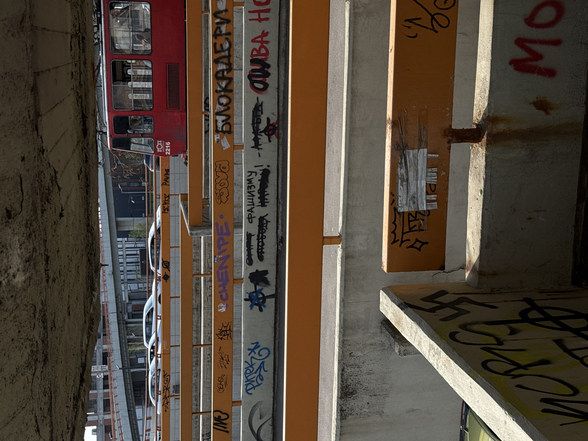 The image captures an upward perspective from beneath an elevated urban structure, likely a bridge or overpass, revealing layers of concrete and yellow-orange metal beams. The scene is rich with textures and human marks, particularly extensive graffiti.
On the left, a red tram with multiple windows is visible on an upper track, moving horizontally across the frame (when viewed upright). Inside, several passengers can be faintly seen. The tram bears the markings "ГОП" (GOP) and the number "2216". Below the tram, a street level is partially visible with parked white cars and other urban buildings in the background.
The concrete pillars and metal beams forming the underside of the elevated structure are heavily adorned with graffiti, predominantly in Cyrillic script and various tags. Notable inscriptions include "БЛОКАДЕРИ" (Blockaders), "CHENIPE", and the phrase "АНТИФАШИЗМУ!" (Anti-Fascism!). Other fragmented words like "СЛОБОДА" (Freedom), "НАРОДУ" (To the people), and "МО" are present. Specific tags like "РАШКА" (Rashka), "PAVKS", and "PERKE" are also visible. A distinct swastika symbol is prominently spray-painted in black on a concrete ledge in the lower right portion of the frame.
The lighting suggests a clear daytime, possibly late morning or afternoon, with strong shadows cast by the structural elements. Rust stains are visible on some metal joints, adding to the gritty, industrial aesthetic. The overall impression is one of a busy, lived-in urban environment, characteristic of Belgrade, Serbia, where such trams and graffiti are common features.