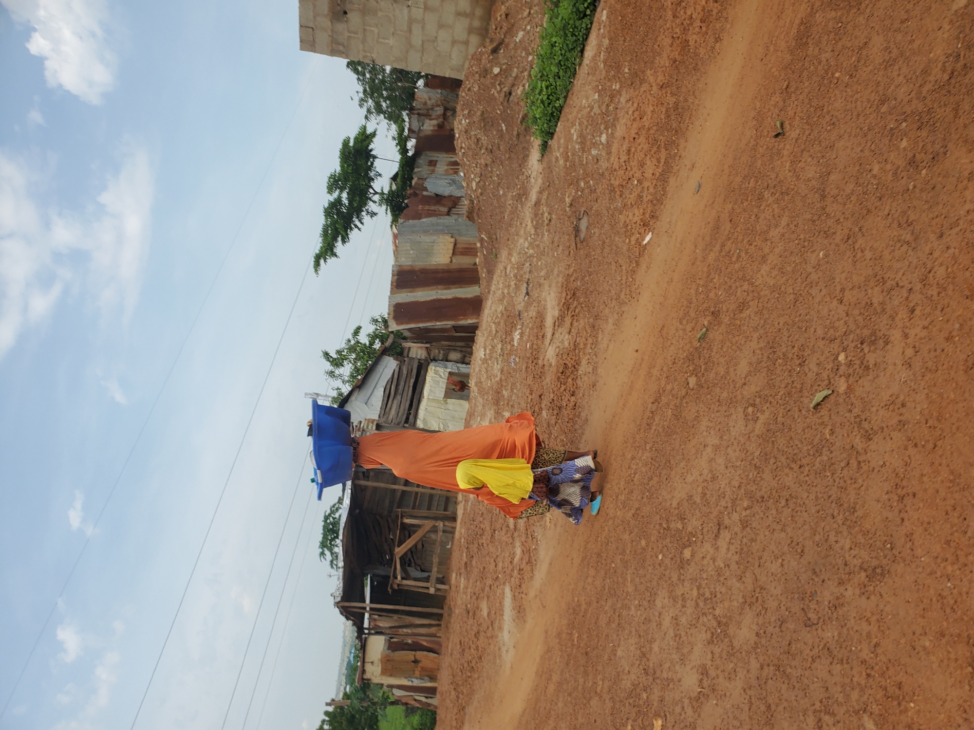 A woman and a child walk on a dirt road in a village. The woman is wearing an orange headscarf and dress, carrying a blue basin on her head. The child, wearing a yellow headscarf and patterned dress, walks beside her. In the background, there are several simple structures made of corrugated metal and wood, characteristic of a rural or developing area. The ground is reddish-brown dirt, and the sky is partly cloudy.