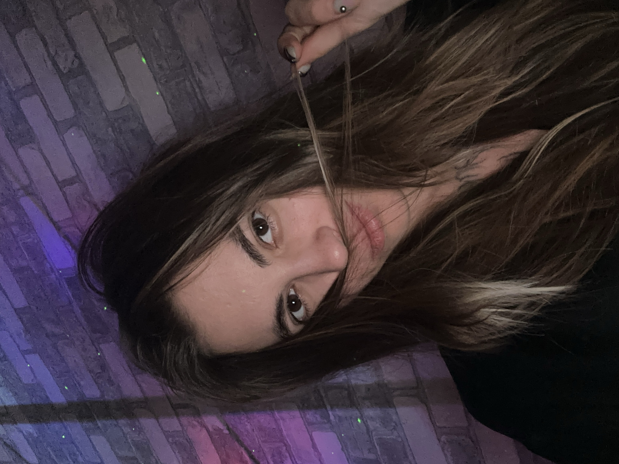 A young woman is taking a selfie against a brick wall illuminated by purple and blue lights, with small green dots visible on the wall. She has long, brown hair, and is pulling a strand of hair across her face. She is wearing a black top. A tattoo is visible on her neck. She has a nose ring and a stud earring. The scene is dark, with the lighting suggesting an indoor environment. No specific landmarks of Moscow, Russia, are visible.