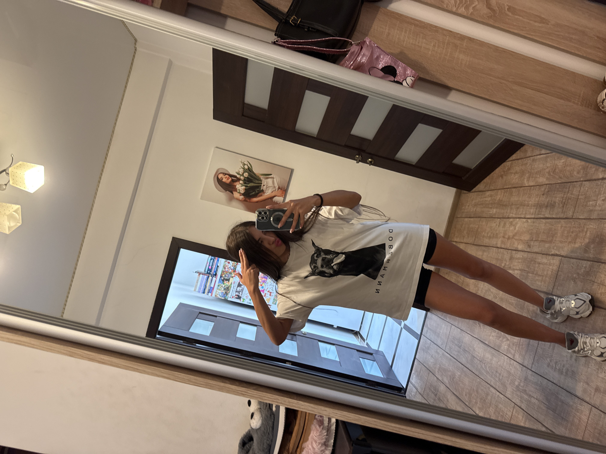 A young woman with long dark hair is taking a mirror selfie in what appears to be a residential hallway in Chișinău, Moldova. She is wearing a white t-shirt with a graphic of a Doberman and black shorts, paired with grey and white athletic shoes. She is holding a phone in her right hand and making a "peace" sign with her left hand near her forehead. In the reflection of the mirror, two doors are visible, one with a white panel and the other a dark wood with frosted glass inserts. To the left of the woman, a stack of grey fuzzy slippers and pink fuzzy slippers are visible on a shelf. On the wall above a door, a framed picture of a woman holding tulips is hanging. The floor is made of light brown wooden planks. The overall environment suggests a domestic interior setting.
