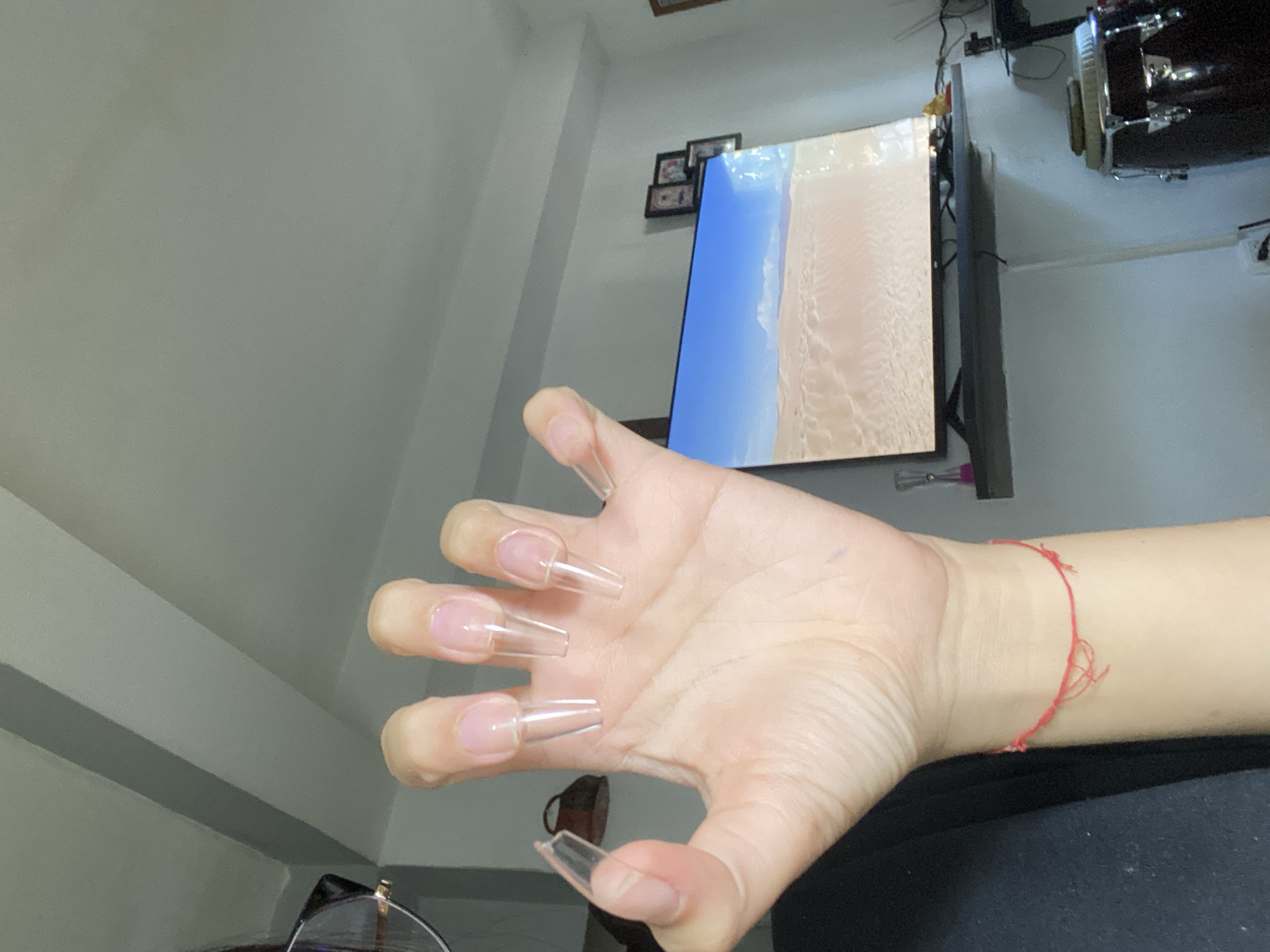 A person's hand is in the foreground, with long, clear, acrylic nails. The background includes a television displaying a beach scene, framed photos, a drum set, and a portion of a pair of glasses. The location cannot be determined from the image.