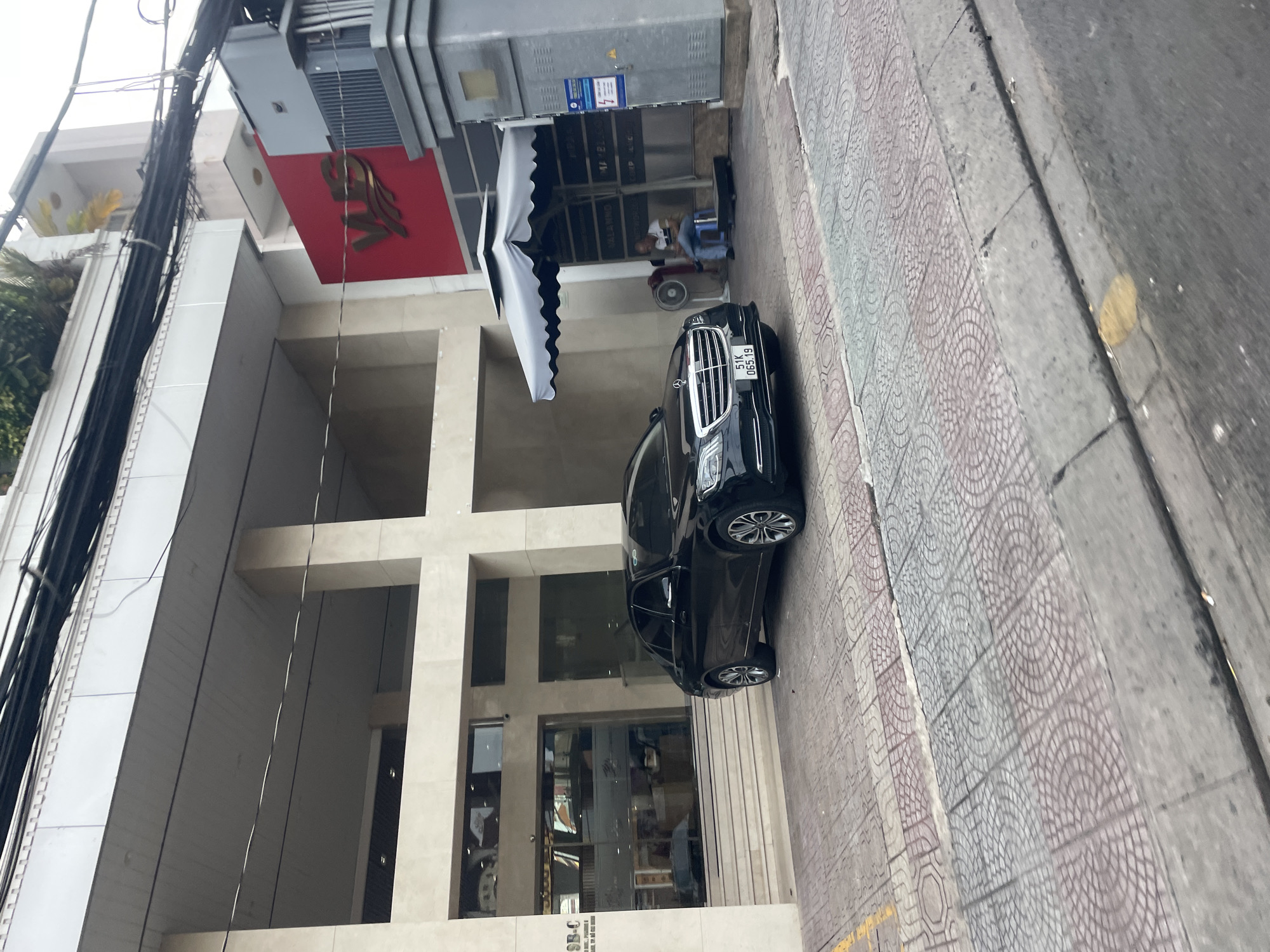 The image captures a black Mercedes-Benz S-Class sedan parked on a patterned tiled driveway in front of a modern building. The building has a beige facade with large glass windows and a prominent red sign with gold lettering that reads "VUS." The car's license plate is visible and reads "51K 065.19". To the right of the car, a man in a white shirt and blue jeans is sitting on a blue stool under a gray and white umbrella next to an outdoor fan and an electrical utility box. The scene is on a street in Thủ Đức, Vietnam, as indicated by the "VUS" sign, a common English language center chain in Vietnam. The environment suggests an urban setting with overhead electrical wires and a sidewalk in the foreground.