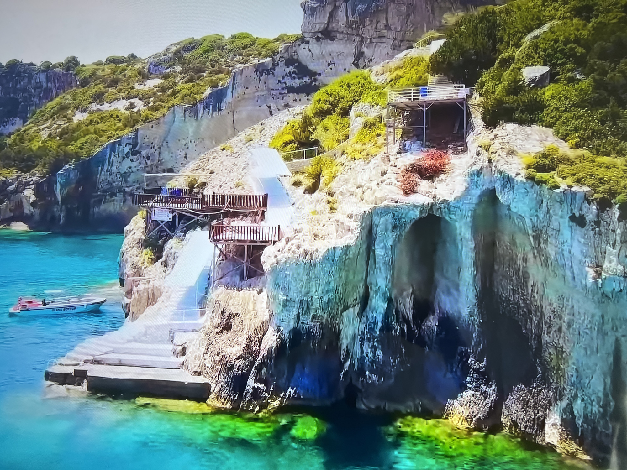 The image presents a coastal scene featuring rocky cliffs and a turquoise sea. Situated along the cliffs are several structures, including wooden platforms and staircases leading down to the water. A small boat, labeled "Glass Bottom," is visible in the water near a platform. The cliffs are covered in vegetation. The water displays varying shades of green near the shore, transitioning to deeper blue hues further out. The presence of man-made structures and a boat suggests recreational or tourist activity in a naturally picturesque location. I am unable to identify the precise location in Basingstoke and Deane, United Kingdom.