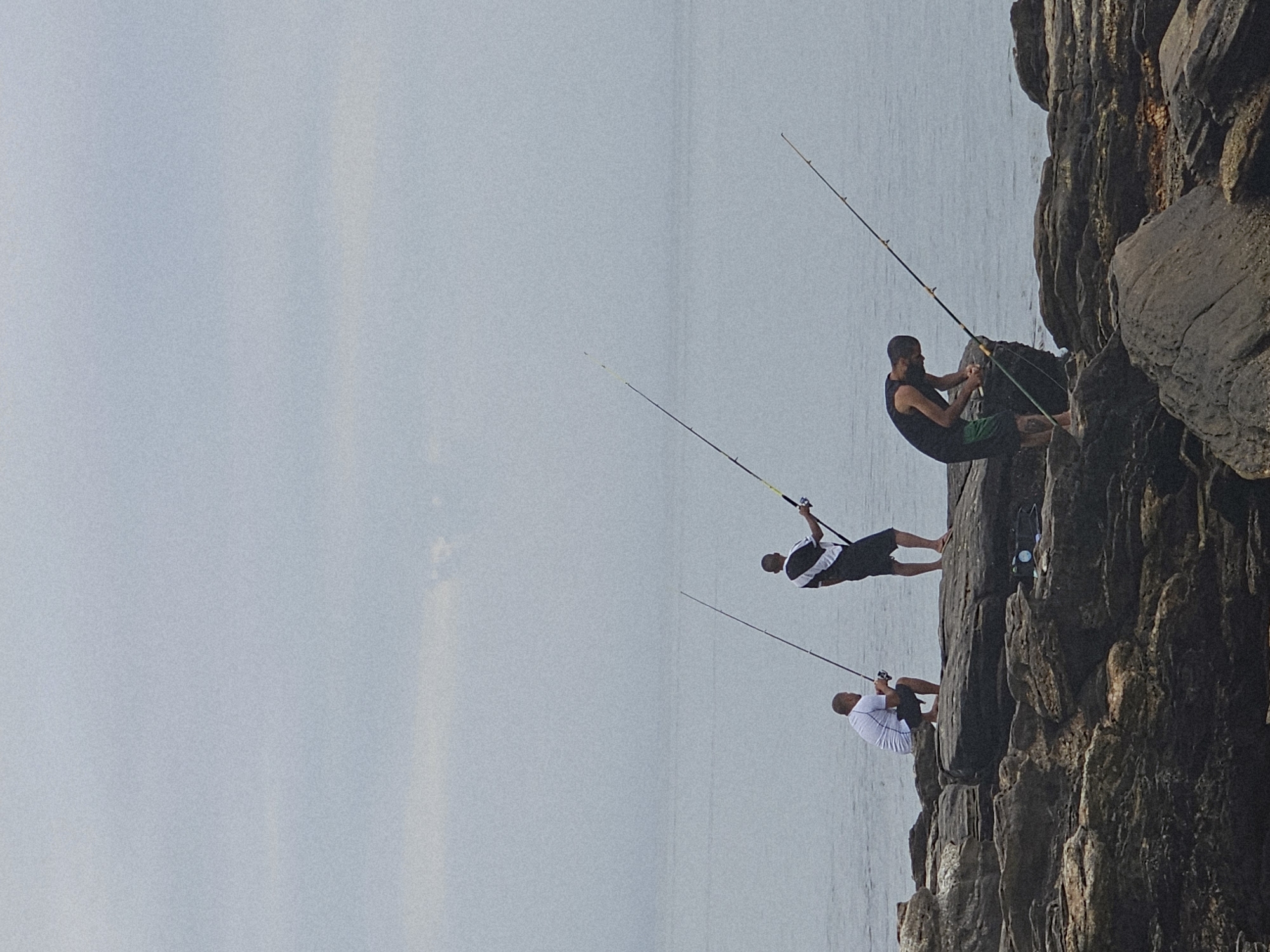 The image depicts three men fishing from a rocky cliff face, likely near the ocean, in Armação dos Búzios, Brazil. The men are spaced vertically along the cliff, each holding a fishing rod. The first man, nearest to the top, wears a black sleeveless shirt and green shorts. The second man wears a black and white shirt and black shorts. The third man, at the bottom, wears a white shirt and black shorts. The backdrop includes a grey sky and calm ocean waters. The cliff is composed of dark, jagged rocks.