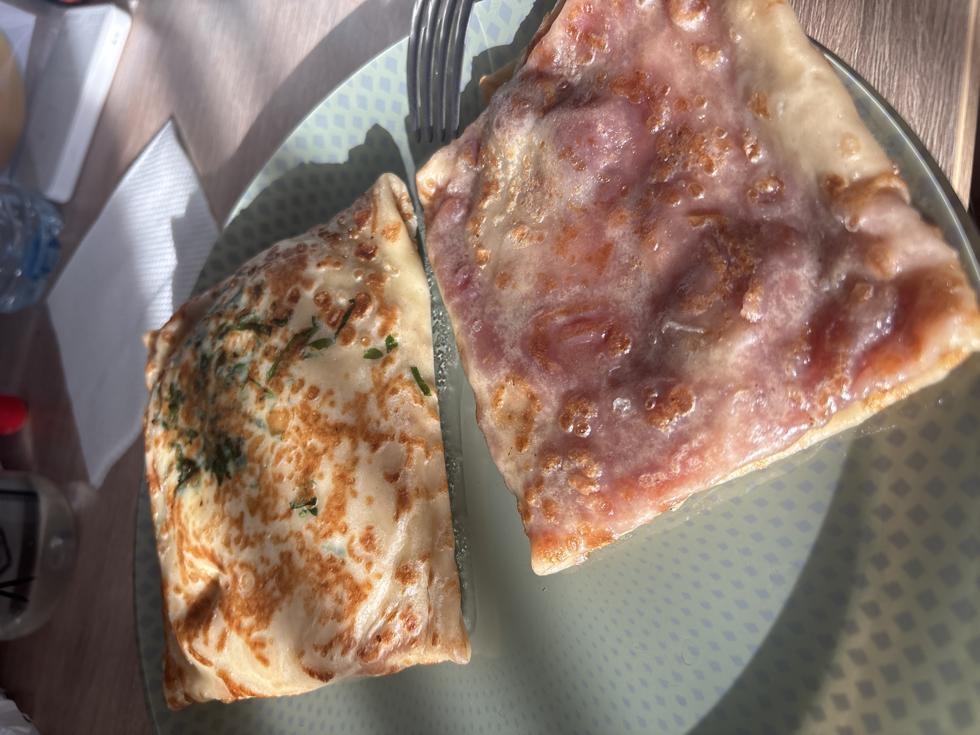 The image shows a plate with two folded pancakes or crepes, possibly in Ladozhskii Trudposelok, Russia, given the prompt. A fork rests between them. The crepe on the left has a golden-brown surface speckled with green herbs, while the crepe on the right is lighter with a pinkish-red filling or topping. The plate has a light-colored surface with a repeating square pattern. A bottle and a napkin are visible in the background, along with a wooden surface. The lighting is bright, with strong shadows.
