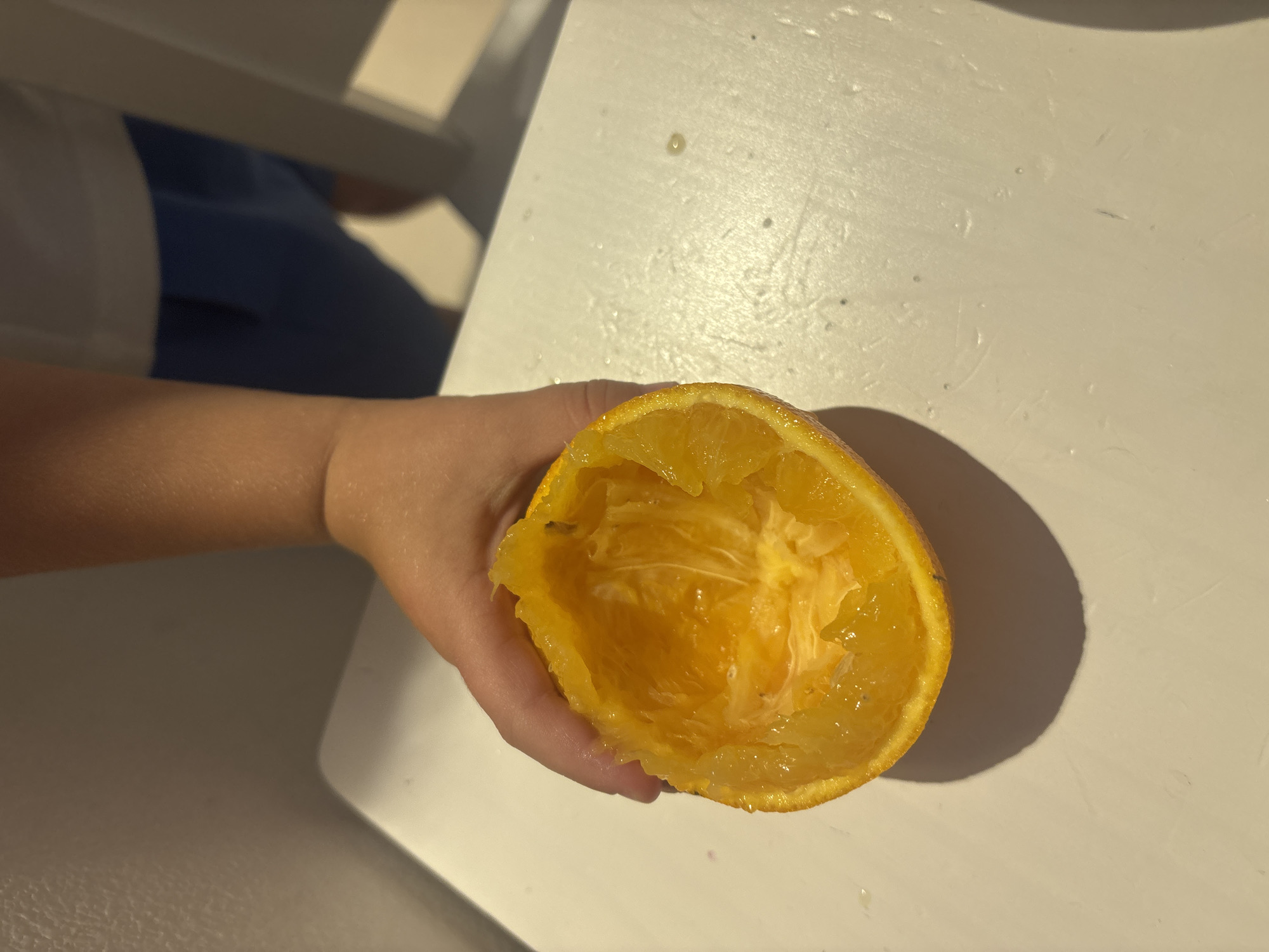 In a bright indoor environment, possibly a kitchen, a person's hand holds a squeezed orange peel. The orange peel is hollowed out and glistening with residual juice. The background is out of focus, showing a white surface, possibly a table, and parts of clothing, including blue shorts and a white shirt. The lighting suggests daytime. There is no information to determine the location in Dubai, United Arab Emirates.