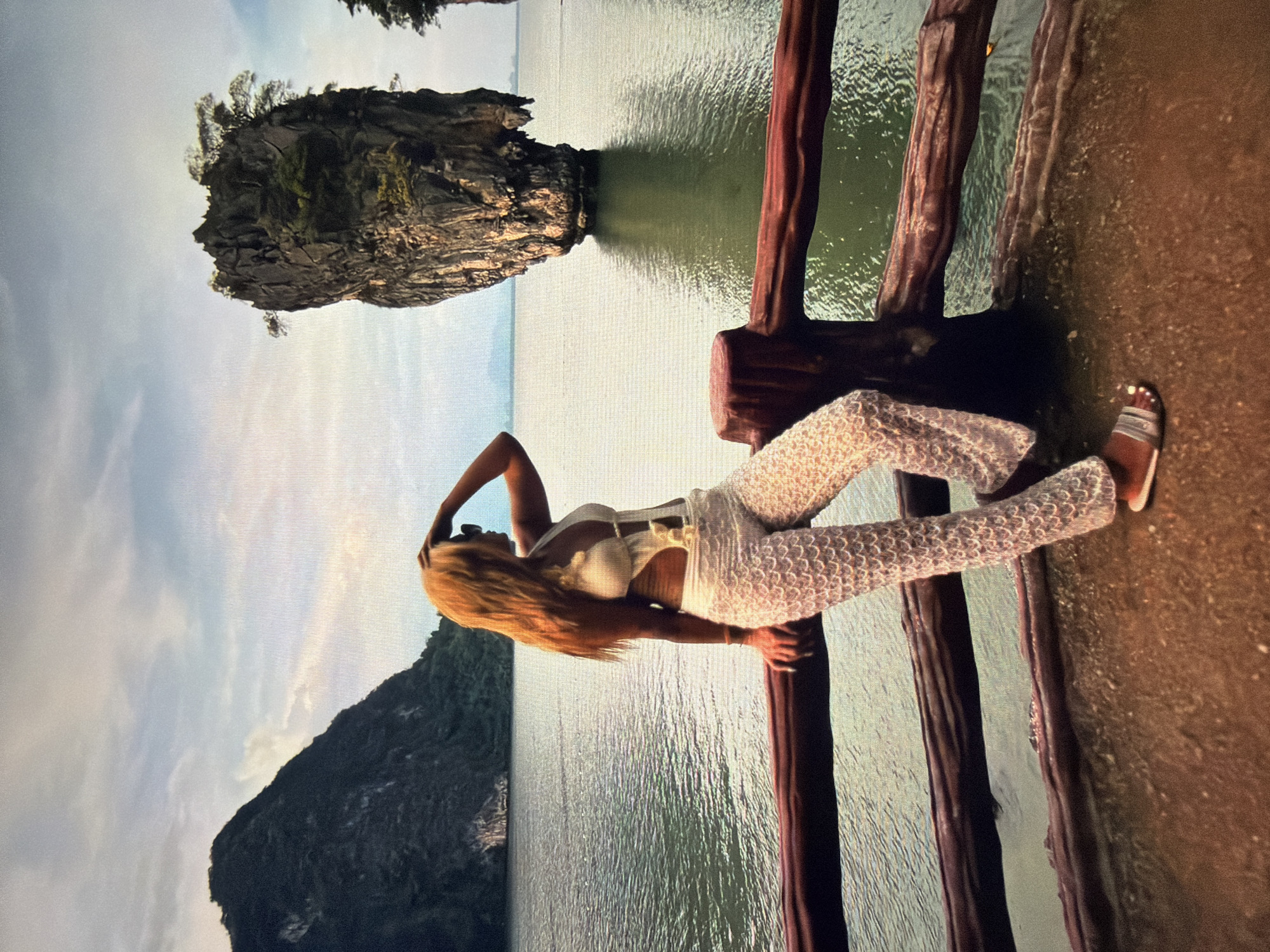 A woman with long blonde hair, wearing a white sleeveless top and light-colored patterned pants, stands on what appears to be a wooden pier or walkway. She is posing with her left hand resting near her head and her right hand holding onto a wooden railing. Her attire includes sandals.
The setting is a picturesque coastal landscape, likely a bay or sea, characterized by large, imposing limestone karsts or islands covered in lush green vegetation. The water is calm, reflecting the cloudy sky. One prominent karst formation is visible in the background, appearing to jut out from the water. Another large landmass or karst is visible further in the background to the left. The sky is partly cloudy, suggesting it is daytime, possibly late afternoon or early morning due to the soft lighting. The wooden railing the woman is leaning on features sturdy, rustic posts. There is no visible text in the image.