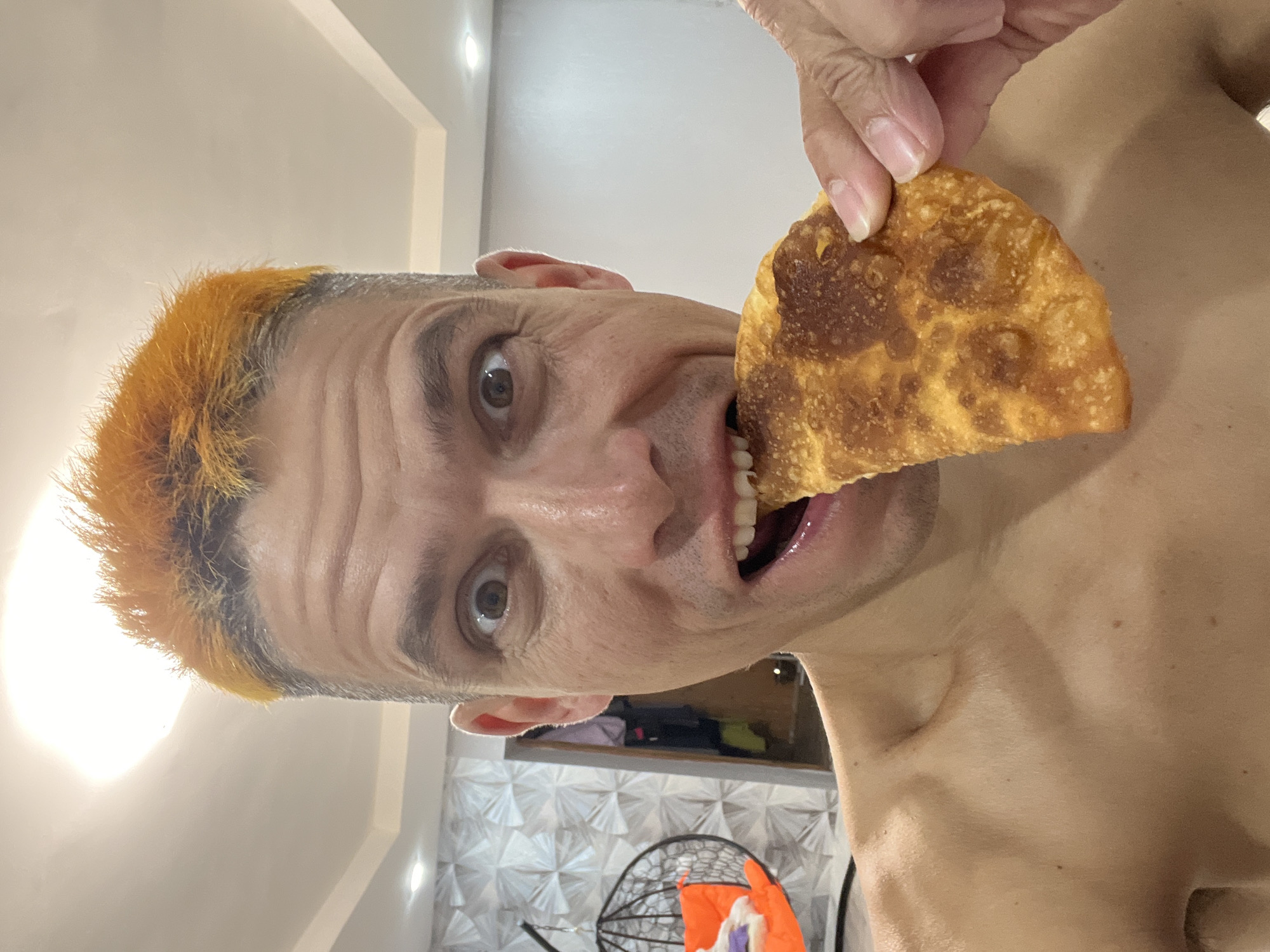A shirtless man with bright orange hair is taking a bite out of a golden-brown, fried pastry. His eyes are wide and looking directly at the camera. He is holding the pastry with his right hand. The background appears to be an indoor setting with textured walls and a ceiling light. There is a hanging chair with an orange cushion visible in the background to the left. The scene does not provide any specific indicators of location within the city of Chaikovsky, Russia.