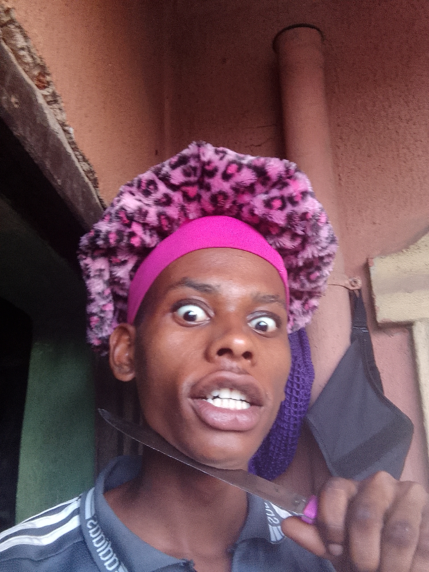 A person with brown skin and wearing a pink headband, a pink and black fuzzy hat, and a gray polo shirt with white stripes is holding a knife to their neck. Their eyes are wide and their mouth is open, seemingly in a comical expression. Behind the person is a wall with a pipe and other objects. The location is unknown.