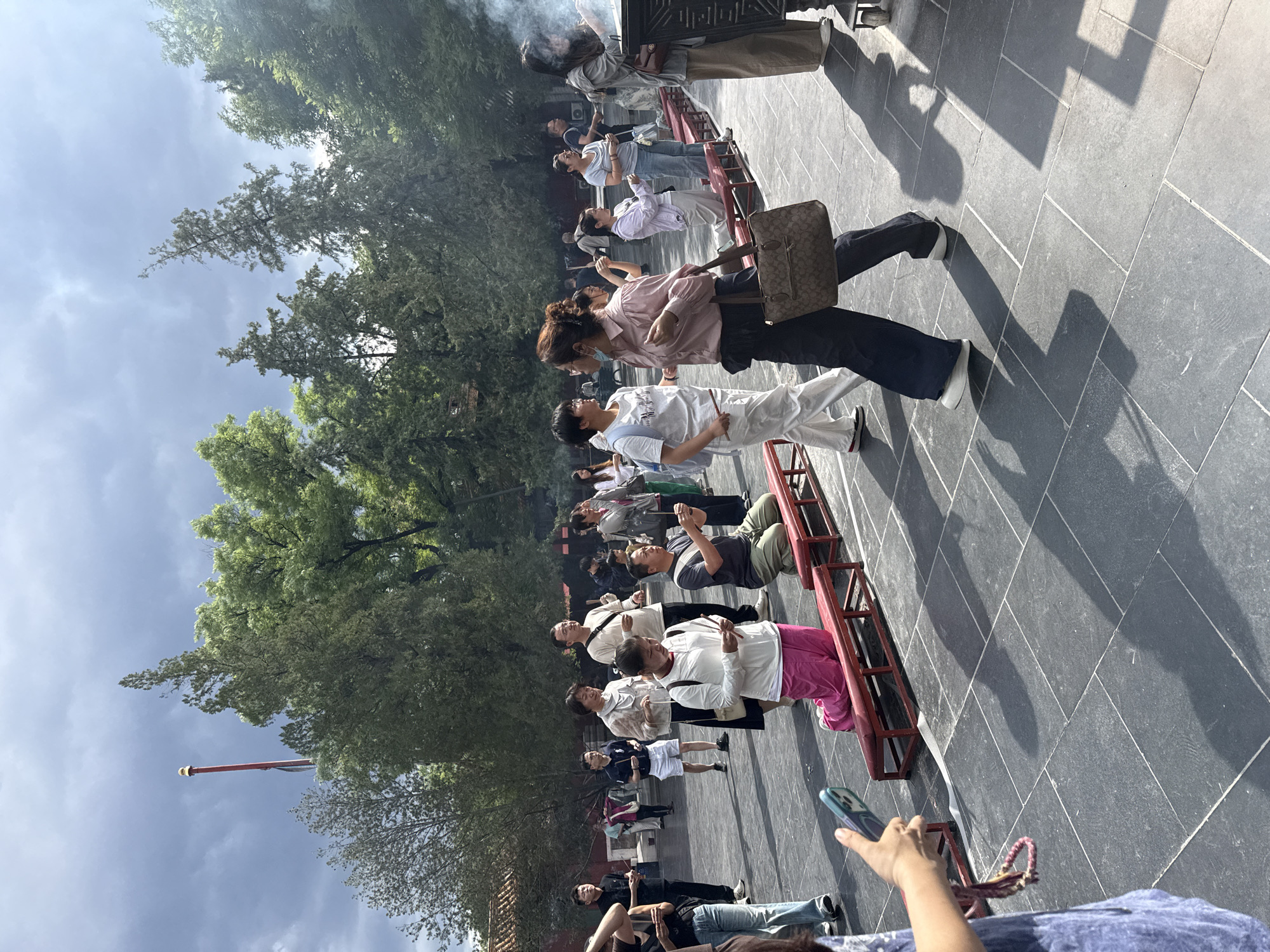 The image depicts a group of people standing in a line outdoors, facing a structure with red supports. Smoke is visible in the upper left of the image, suggesting incense or burning offerings. The people are dressed in casual clothing, and some appear to be holding sticks, possibly incense. The ground is paved with dark grey tiles arranged in a grid pattern, and shadows are cast by the people and the structure, indicating it is daytime with sunlight. Trees with green foliage are visible in the background, suggesting an outdoor setting, possibly a temple or historical site, consistent with the Dongcheng District, China location context. The mood appears calm and perhaps reverent, given the likely context of incense burning. No visible text is present in the image.