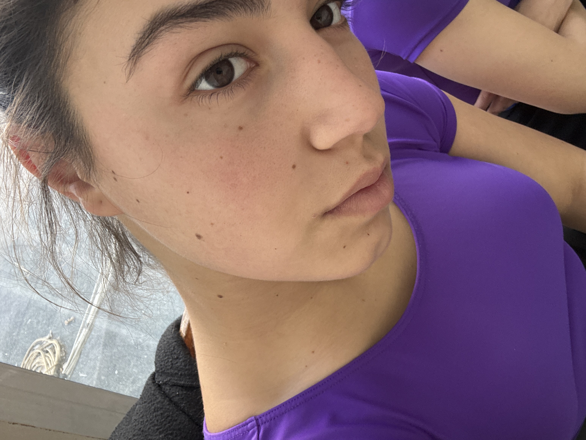 The image is a close-up portrait of a young woman, likely a selfie, taken indoors in Sofia, Bulgaria. The subject has fair skin, dark brown eyes, and prominent dark eyebrows. Her dark hair is pulled back, with some loose strands framing her face and ear. Several small moles or freckles are visible across her cheeks, nose, and neck. She has a neutral to slightly serious expression, looking directly into the camera.
She is wearing a bright, solid purple top with a rounded neckline, possibly athletic wear. A dark, possibly black, garment is visible over her left shoulder, suggesting she may be wearing a jacket or another layer. In the background, a window provides a blurred view of the outside, revealing some indistinct objects like a woven natural fiber item and thin white strands. Part of another person or a reflection is visible in the upper right, also wearing a matching purple top, suggesting shared attire or an activity involving multiple participants. The lighting appears to be natural daylight. No visible text is present in the image.