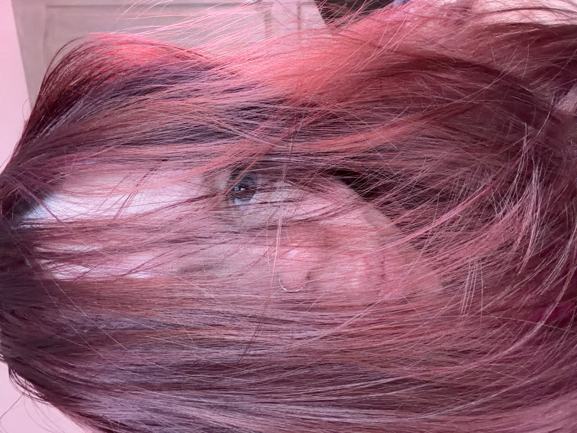 A close-up photograph features a person with their face partially obscured by long, wavy, reddish-purple hair. An eye is visible through the hair, gazing forward. A portion of the ear and skin of the face are also visible. The background is out of focus and appears to be a light pink or peach color. The image composition is tight, focusing primarily on the hair and the visible portions of the face. There is no indication of the location being in Chaikovsky, Russia.