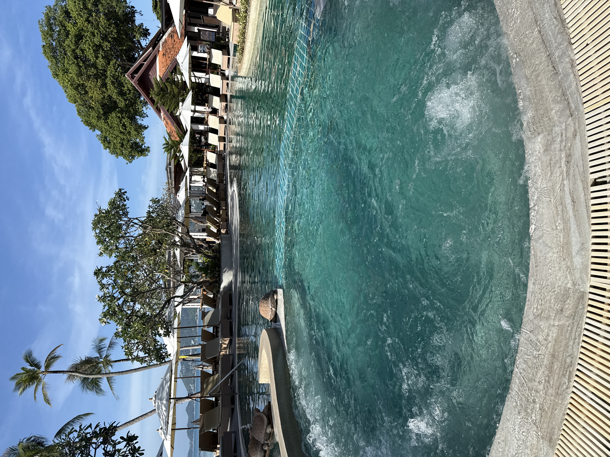 An aerial view shows a resort swimming pool with turquoise water, palm trees, and lounge chairs under umbrellas in Ko Samui District, Thailand. Two turtle-shaped statues are visible in the foreground. A tiled wall separates the pool into sections, and jets create bubbles in the water. In the background, resort buildings with tiled roofs are visible. The sky is clear and blue.