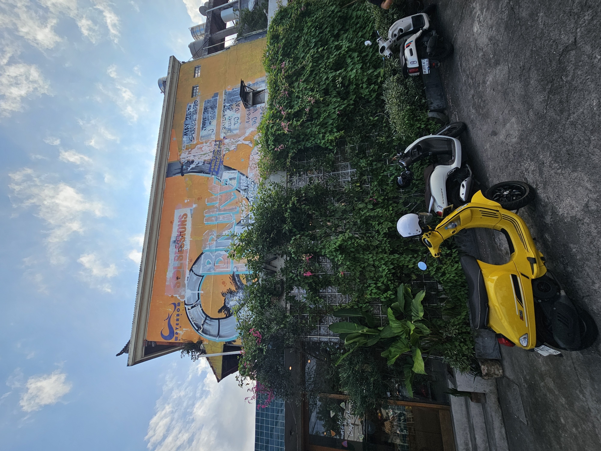 The image depicts an outdoor scene in North Kuta, Indonesia. A tall, yellow-orange building with faded graffiti and advertisements covers most of the left side of the frame. Lush green vines and plants climb the building's walls, partially obscuring the architectural details.
Several motor scooters, including a bright yellow one with a white helmet, are parked in a line on the gray concrete pavement next to the building. The scooter furthest from the building appears to have its license plate visible.
Above the building, a blue sky with scattered clouds is visible. The scene suggests a commercial area, possibly a café or shop, given the building's signage and the parked scooters. There is no direct depiction of people in the image.