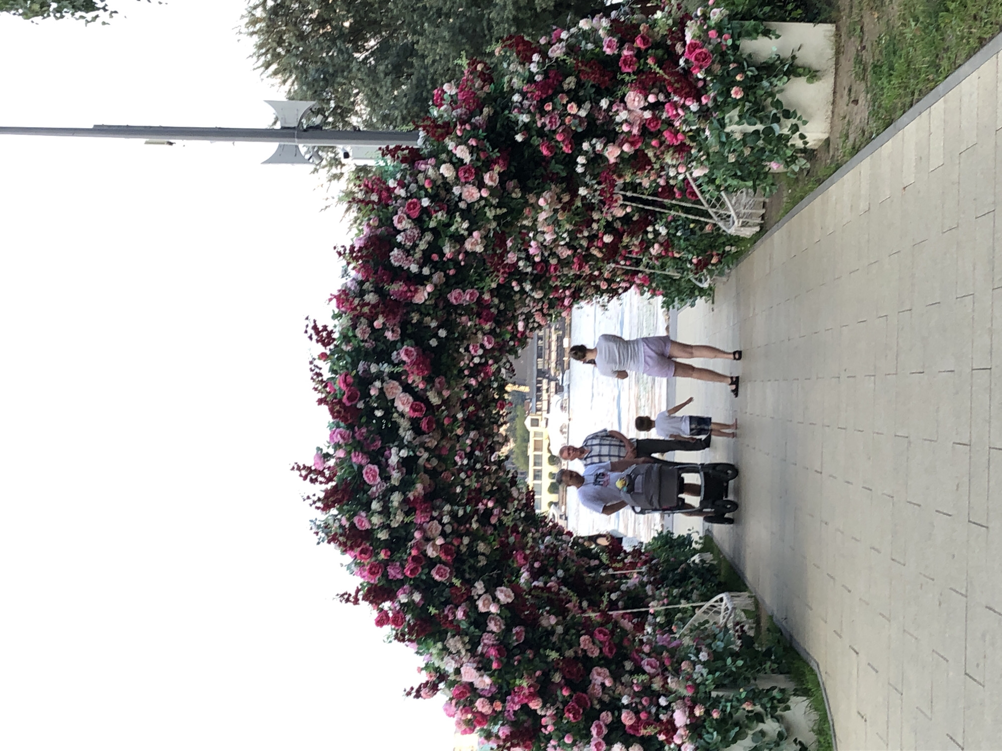 The image depicts a floral archway constructed with numerous pink and red roses and other greenery, spanning across a paved walkway. On the walkway, there are four people. A man and a woman are standing near a baby stroller, facing away from the camera. A young boy is walking towards the floral archway, also facing away from the camera. Another man is standing to the left of the baby stroller, facing towards the floral archway. In the background, there is a body of water with boats on it, and buildings can be seen on the opposite shore. A tall pole with some equipment attached is visible to the right of the floral archway. The scene appears to be a public outdoor space, likely a park or waterfront area in the city of Khimki, Russia.