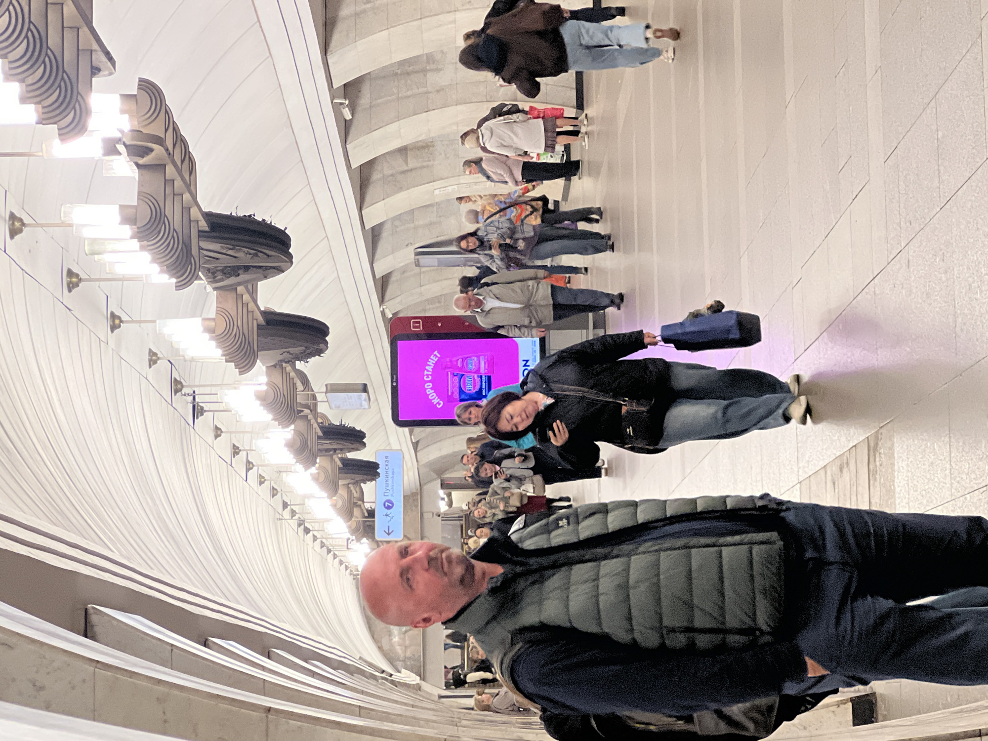 Here is a description of the image:
The image shows a person in a gray puffer vest and dark blue shirt and jeans standing in a subway station. The station has a white, curved ceiling with decorative light fixtures. Several other people are visible in the background, walking or standing. There's a large digital display visible in the background with pink colors on it. A sign with the word "Пушкинская" (Pushkinskaya) is partially visible near the top left of the person.