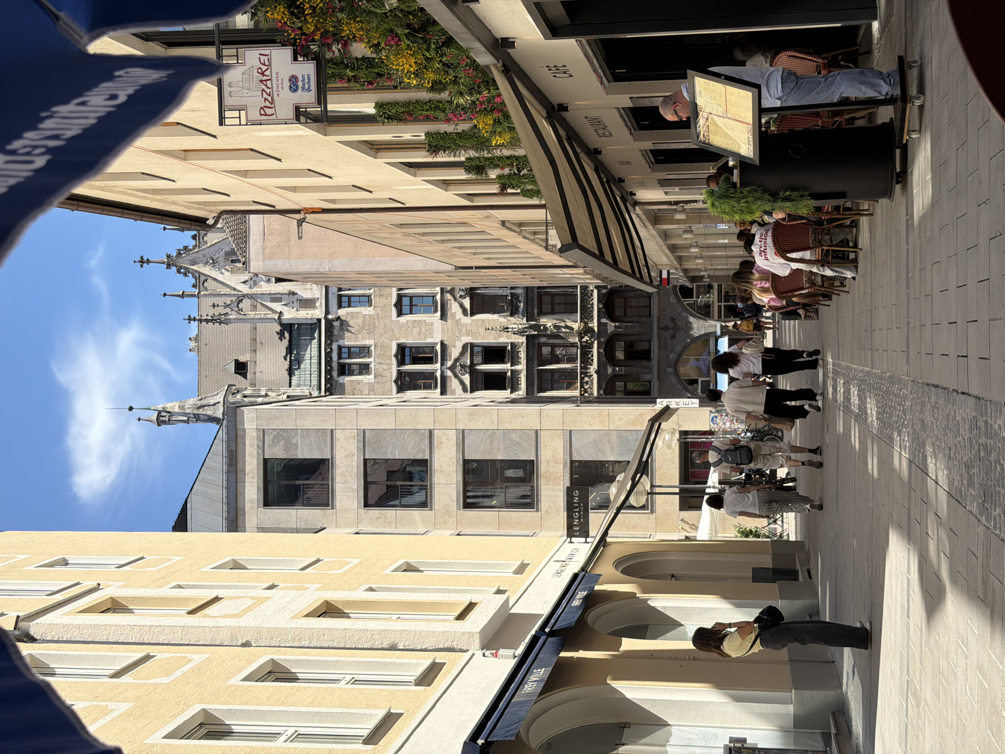 The image depicts a sunny day in a pedestrianized street in Munich, Germany, likely in the city center. The street is lined with multi-story buildings, some with yellow stucco facades and others with stone cladding. Architectural styles vary, with gothic elements visible in the background.
On the left side of the street, there are shops with awnings, one of which is labeled "FREY VILLE." A woman in a yellow top and dark pants is standing near the entrance of a shop, looking at her phone.
The right side of the street features a cafe or restaurant with outdoor seating. Several people are seated at tables, and a man in a blue shirt and jeans is standing by a large menu board. The building on the right has a sign that says "PIZZAREI" and also "CAFE" and "RESTAURANT" are visible on other establishments. Verdant greenery and colorful flowers are arranged on the building facades, adding to the ambiance.
The street itself is paved with cobblestones, and shadows from the buildings and awnings are cast across the ground, indicating the time of day is likely midday or early afternoon. People are strolling, dining, and engaged in various activities, creating a lively urban atmosphere.
