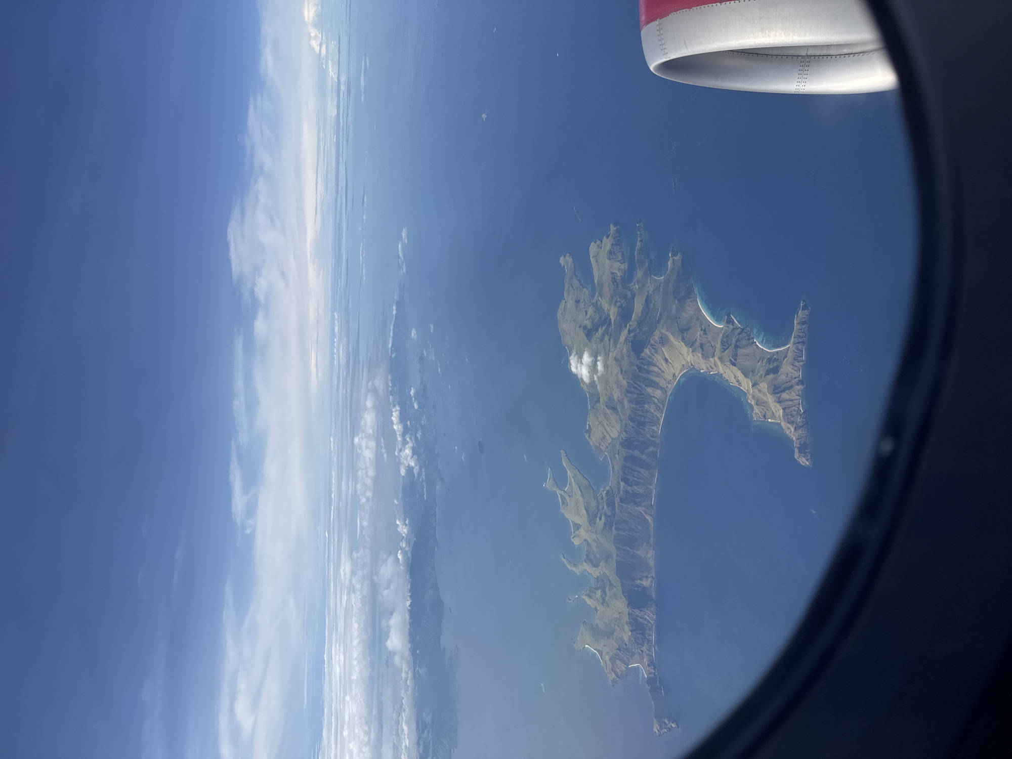 The image shows an aerial view of an island with green hills and white beaches, surrounded by blue ocean. The view is framed by an airplane window with a portion of the engine visible. The sky shows a combination of clear blue and scattered clouds. Location: Unknown, Unknown.