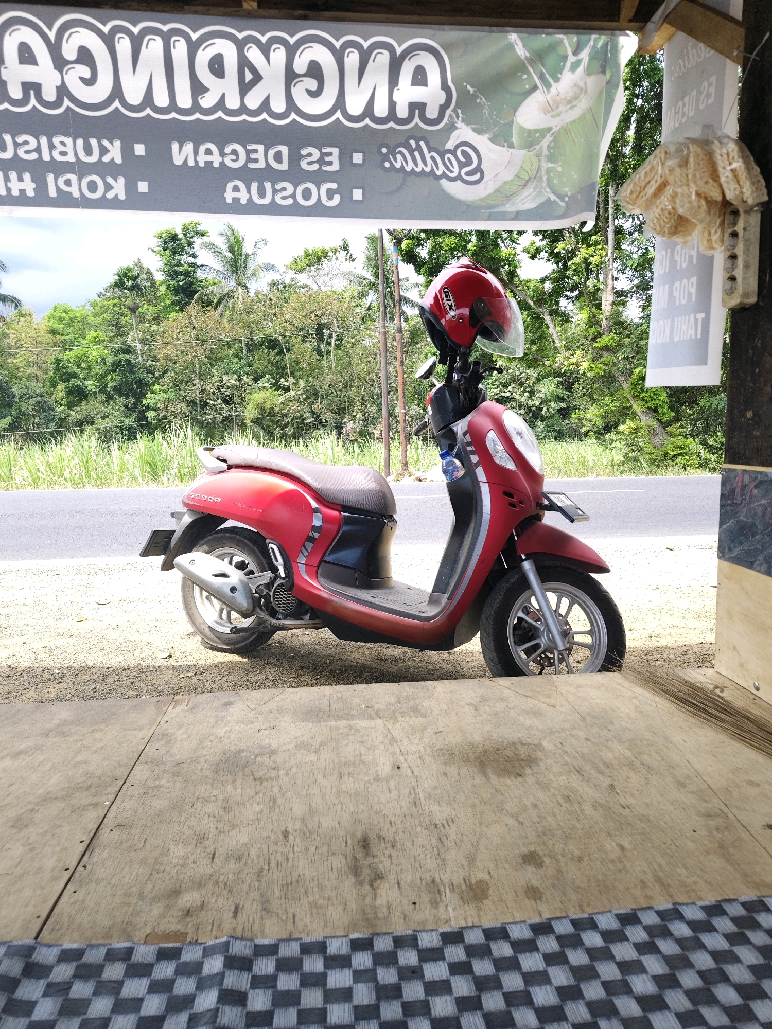 A red Honda Scoopy motorcycle is parked on a gravelly surface next to a road, with a wooden structure and a checkered mat in the foreground. A red helmet rests on the handlebars of the motorcycle. A banner advertising coconuts and drinks hangs above the scene, with text in Indonesian. In the background, lush green trees and foliage are visible. The scene appears to be an outdoor stall or shop in Pondok Dalem, Indonesia, likely a roadside establishment.
