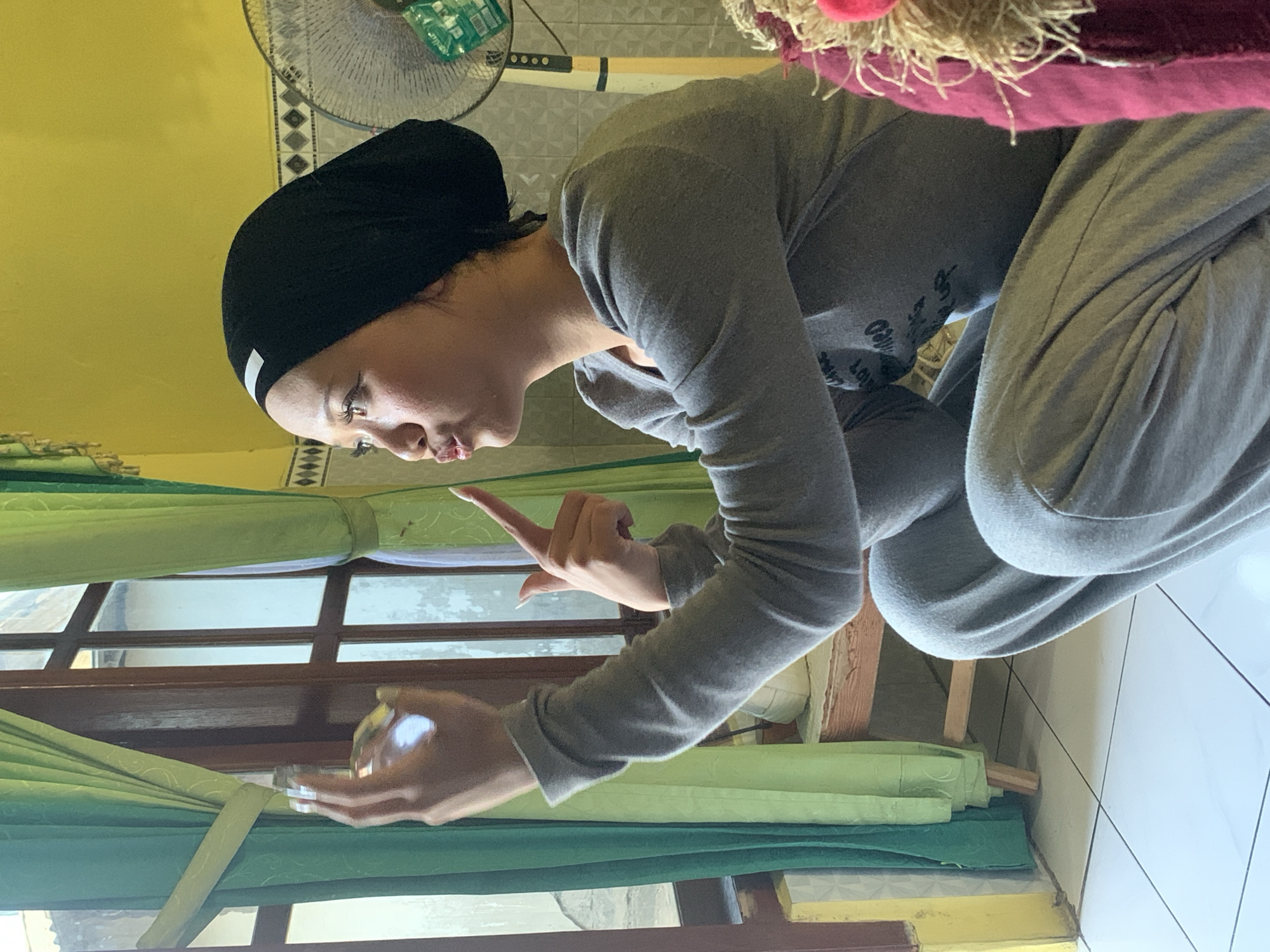 A young woman wearing a black hijab and a grey tracksuit is crouched by a window with green curtains. She is looking to her right, with her mouth pursed, and holding a small, reflective object in her left hand, while pointing with her right index finger. The room has yellow walls and tiled floors. In the background, a fan is visible. The scene appears to be indoors in a residential setting in Jajag, Indonesia.