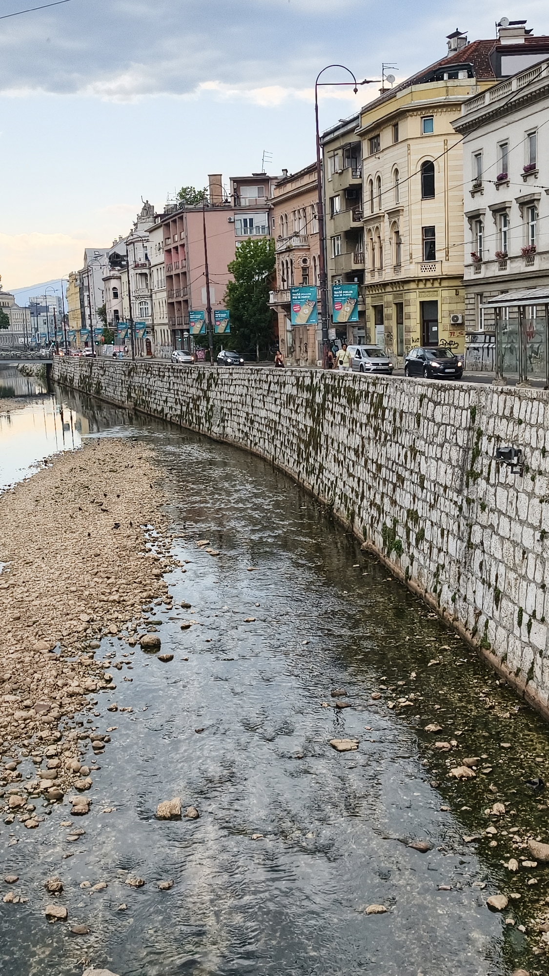 The image shows a view of the Miljacka River in Sarajevo, Bosnia and Herzegovina, with a stone embankment and buildings lining the opposite bank. The riverbed is partially exposed, with rocks and pebbles visible. On the embankment, there is a stone wall with moss and greenery growing on it. Above the wall, a street is lined with multi-story buildings with a mix of architectural styles. Several cars are visible on the street, and pedestrians are walking along the sidewalk. The sky is overcast. The overall impression is an urban scene with historical architecture along a river.