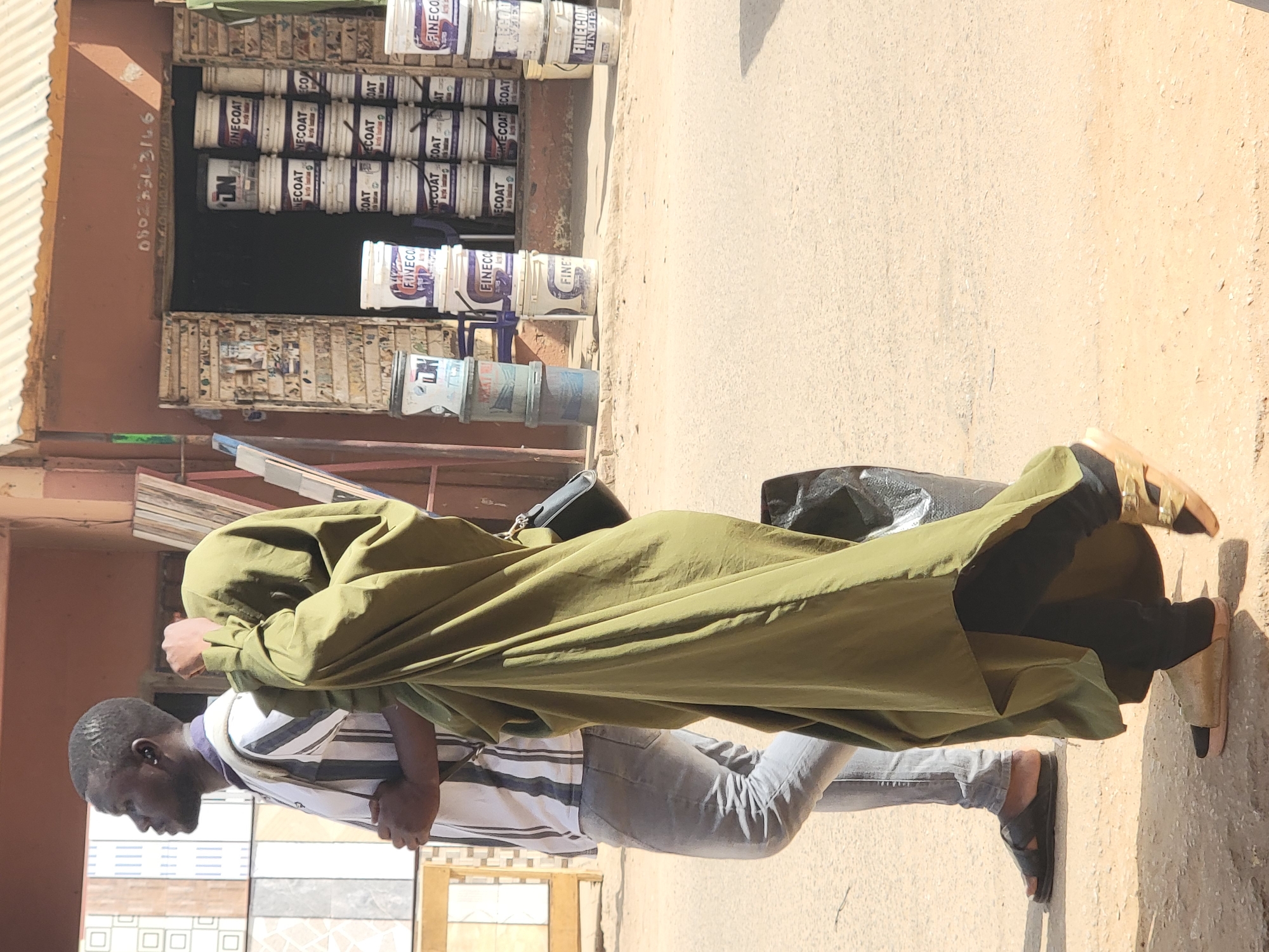 The scene depicts a street in Maiburiji, Nigeria. A person wearing an olive-green, full-length garment walks in front of a building that sells paint and tiles. The individual's face is partially obscured by the hood of the garment. A second person wearing a striped shirt and gray pants walks alongside them. The first person carries a black bag over their shoulder. The background shows a shop stocked with multiple buckets of white "FINECOAT" brand paint. The front of the shop also displays a variety of tiles. A phone number, "0802033146" is written next to the shop. The ground is concrete.