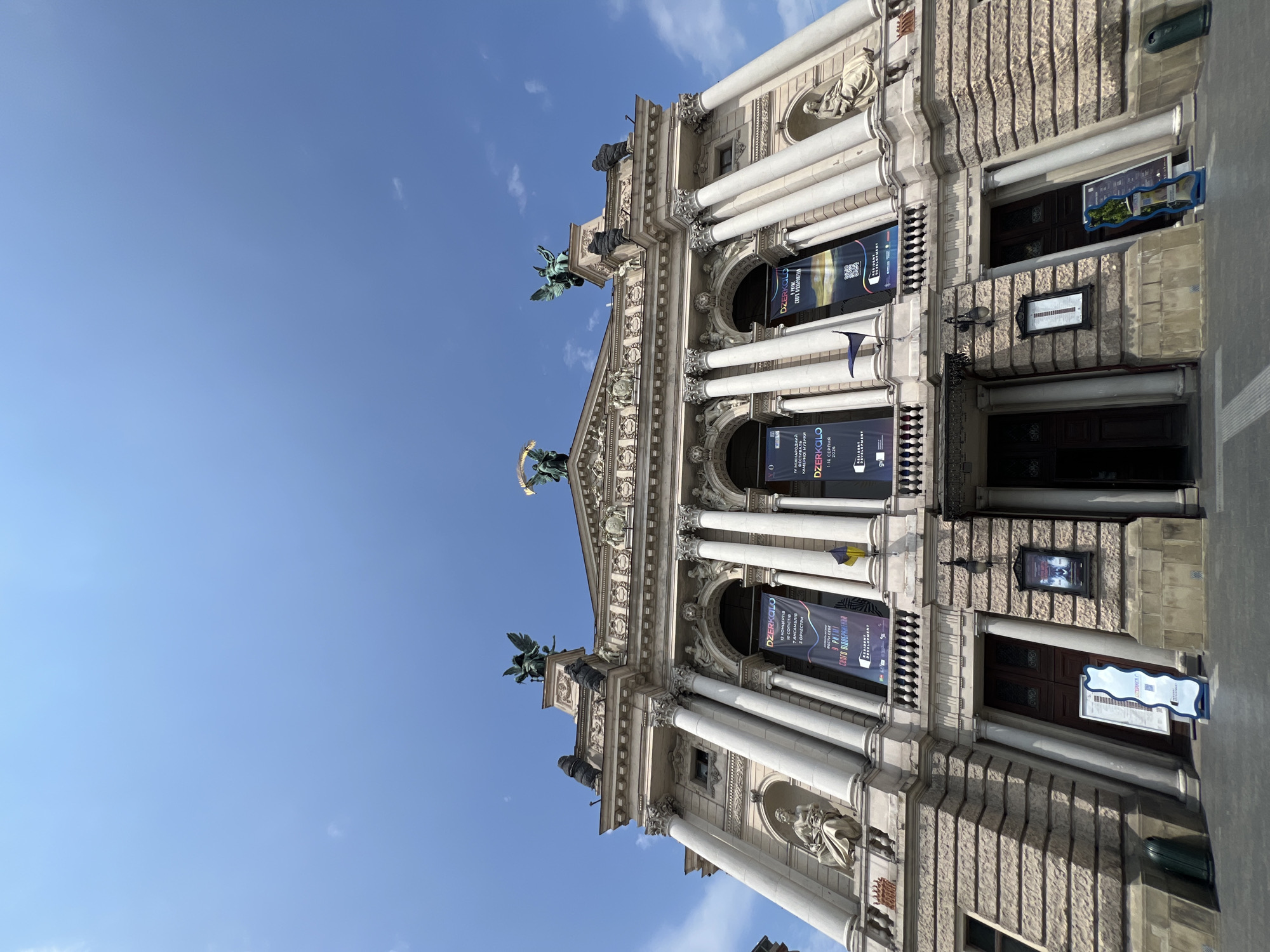 The image shows a low-angle, wide shot of the Lviv Opera House in Lviv, Ukraine, under a clear blue sky with a few wispy clouds. The grand, ornate facade of the building dominates the frame, with its classical columns, sculptures, and decorative stonework. Several large banners, advertising events and featuring text in Ukrainian, hang from the upper levels. A Ukrainian flag is visible near the center banner. In the foreground, a few doors are visible, along with some informational signs and a trash can. The overall impression is one of architectural grandeur and a vibrant cultural setting.