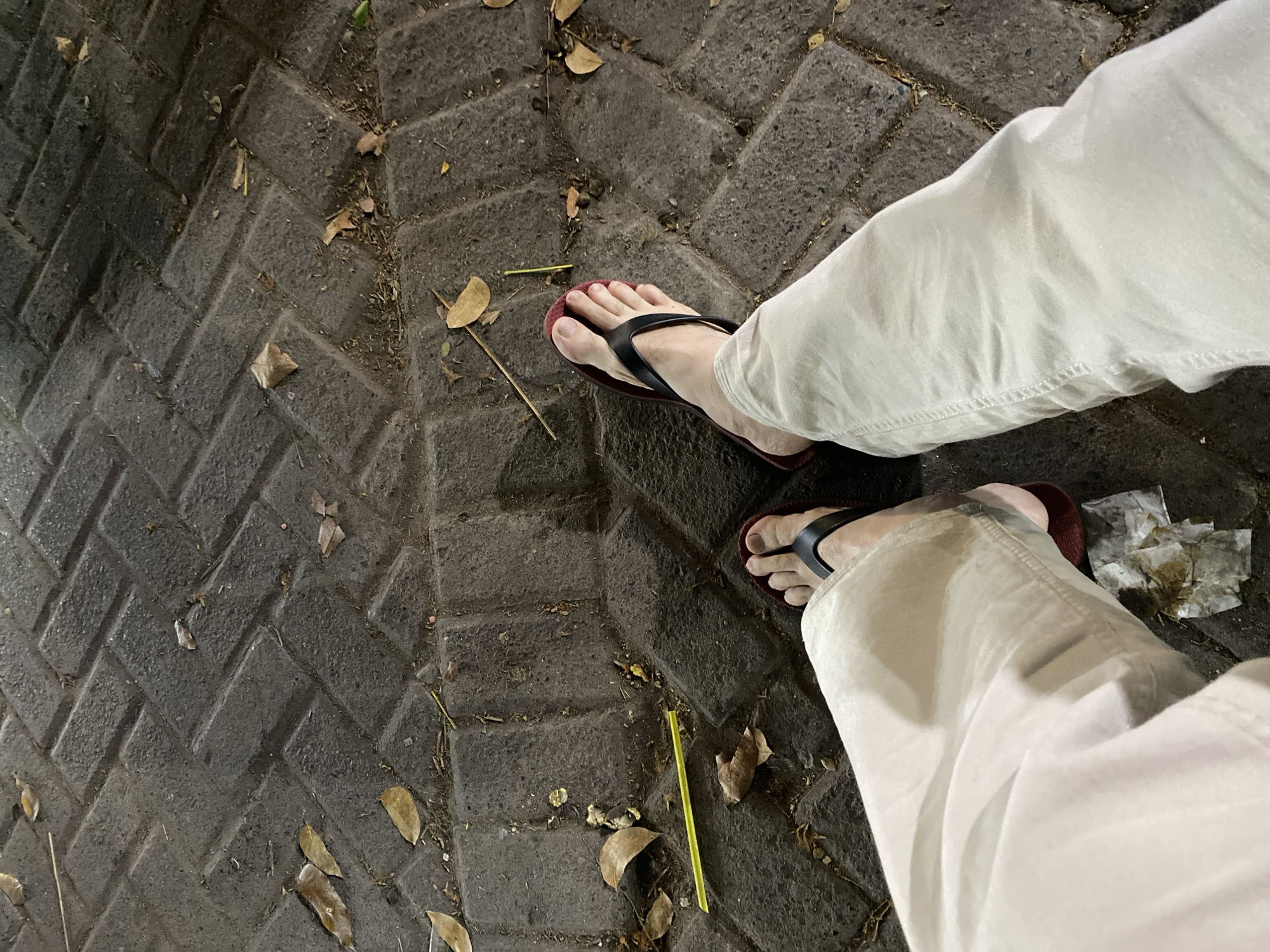 The image shows a close-up view of a person's legs and feet, wearing light-colored pants and flip-flops, standing on a textured paving stone surface. Several dried leaves are scattered on the ground, and a yellow straw is visible near the left leg. The paving stones are arranged in a herringbone pattern and appear to be made of dark concrete. The lighting suggests it is evening or night. The scene is likely a public outdoor area within the city of Banjarmasin, Indonesia, possibly a sidewalk or a public square.
