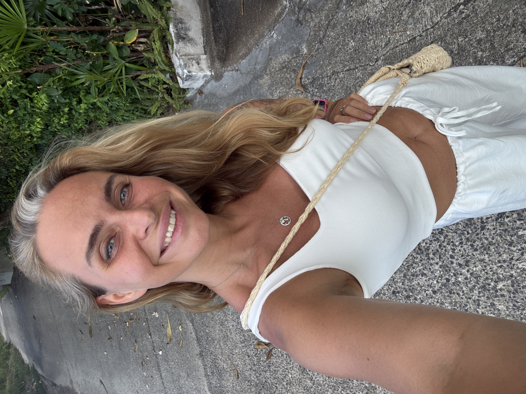 A blonde woman with blue eyes smiles at the camera, taking a selfie. She is wearing a white crop top and white pants, with a small straw crossbody bag. A necklace with a peace sign pendant is visible. She is standing on a paved pathway, with lush green foliage to her right and more foliage and a stone wall in the background. The pathway is wet, suggesting recent rain. The scene appears to be outdoors, possibly in a tropical location given the vegetation. The specific location of Baan Chaweng, Thailand is not discernible from the image itself but is provided as context.
