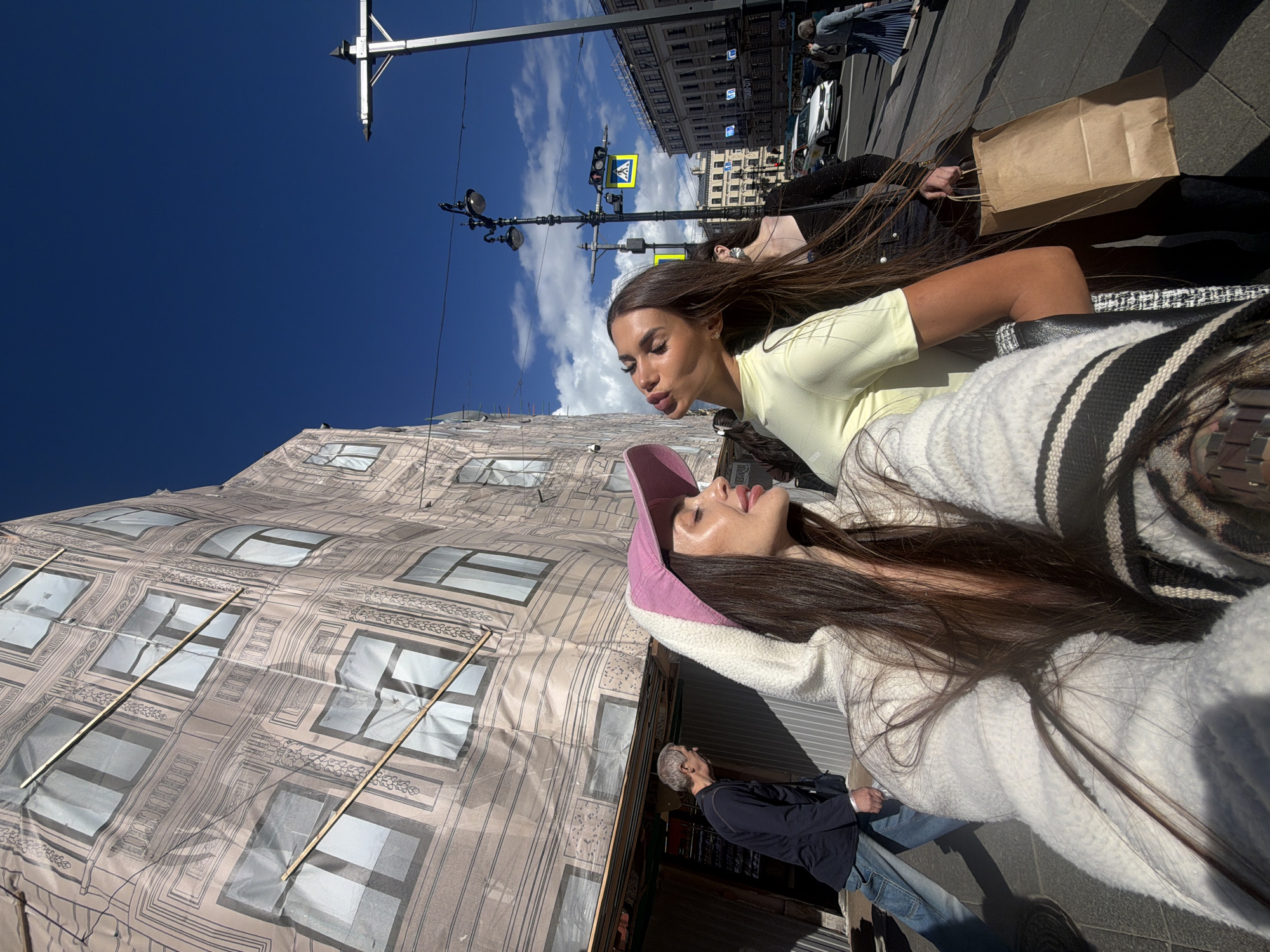 This media file captures an outdoor scene in Saint Petersburg, Russia, under a bright, sunny sky with scattered clouds. The image is taken from a slightly tilted perspective, looking upwards towards a deep blue sky and a building facade covered with a large tarp printed with a pattern of windows and architectural details, suggesting renovation or construction.
In the foreground, two women are the primary subjects. One woman, with long, dark hair, is wearing a light yellow top and leans forward, her face angled upwards with her lips pursed as if blowing a kiss. She appears to be holding a pink object, possibly a mirror. Beside her, another woman, also with long dark hair, is draped in a white textured garment and a black and white striped scarf. Her face is also turned upwards, with her eyes closed and lips parted. Both women seem to be enjoying a casual moment outdoors.
In the background, a street scene unfolds. Pedestrians can be seen walking along a sidewalk, and cars are visible on the road. Traffic lights and street lamps are also present, indicating an urban environment. One person, a man with grey hair, is walking in the mid-ground, looking upwards. A brown paper bag is held by someone out of frame on the right. The sunlight suggests it is daytime, likely late morning or early afternoon, given the brightness and the shadows cast on the ground. The overall mood appears relaxed and cheerful. No visible text is present in the image.