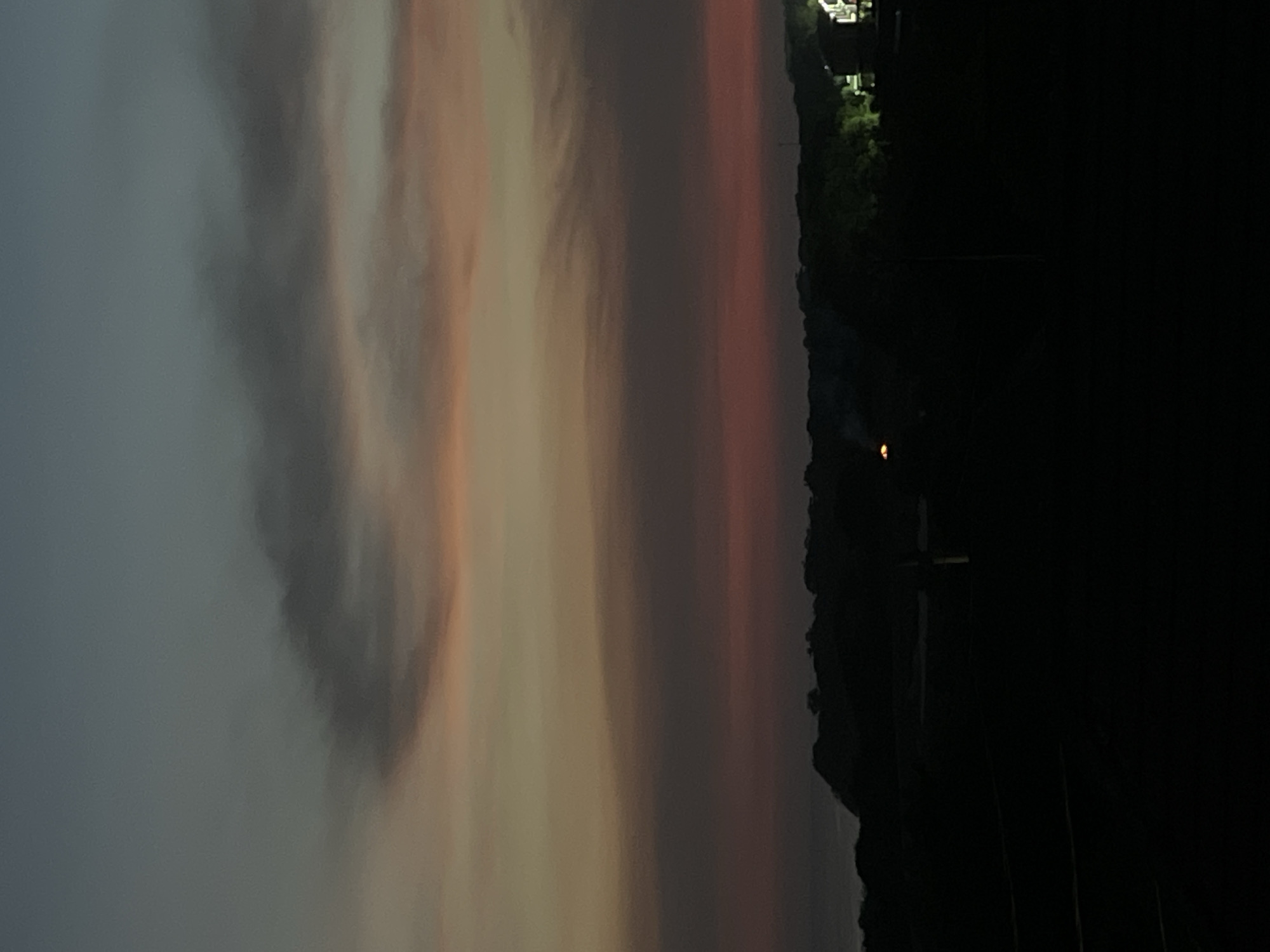 The image depicts a dusky sky transitioning from soft grey to pale orange and pink hues on the left, merging into deeper reddish tones towards the right. A dark, silhouetted treeline defines the horizon on the right side, with what appears to be a cluster of buildings with lights on at the top right. A tall, dark structure resembling a cross or utility pole is visible near the center right, rising against the dark background. The bottom right corner of the image is entirely dark, obscuring any details. The scene gives the impression of a quiet, possibly rural or suburban setting at dusk.