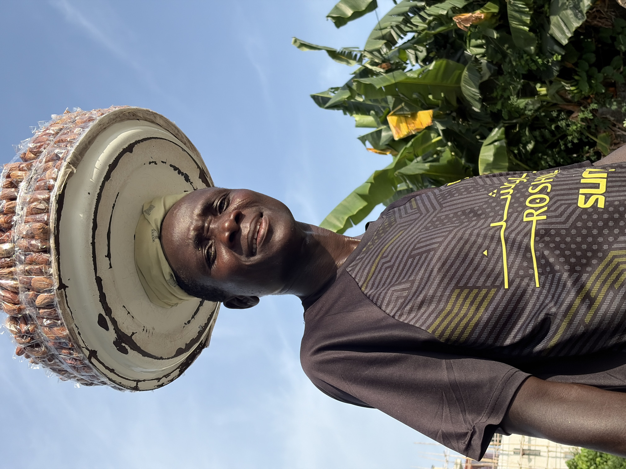 A low-angle shot shows an Igbo man in Igboefon, Nigeria, balancing a large, white, cylindrical container filled with small packets of dates on his head. He is wearing a black t-shirt with a geometric yellow and gray design. The sky is blue, and behind him are banana trees and an unfinished building.