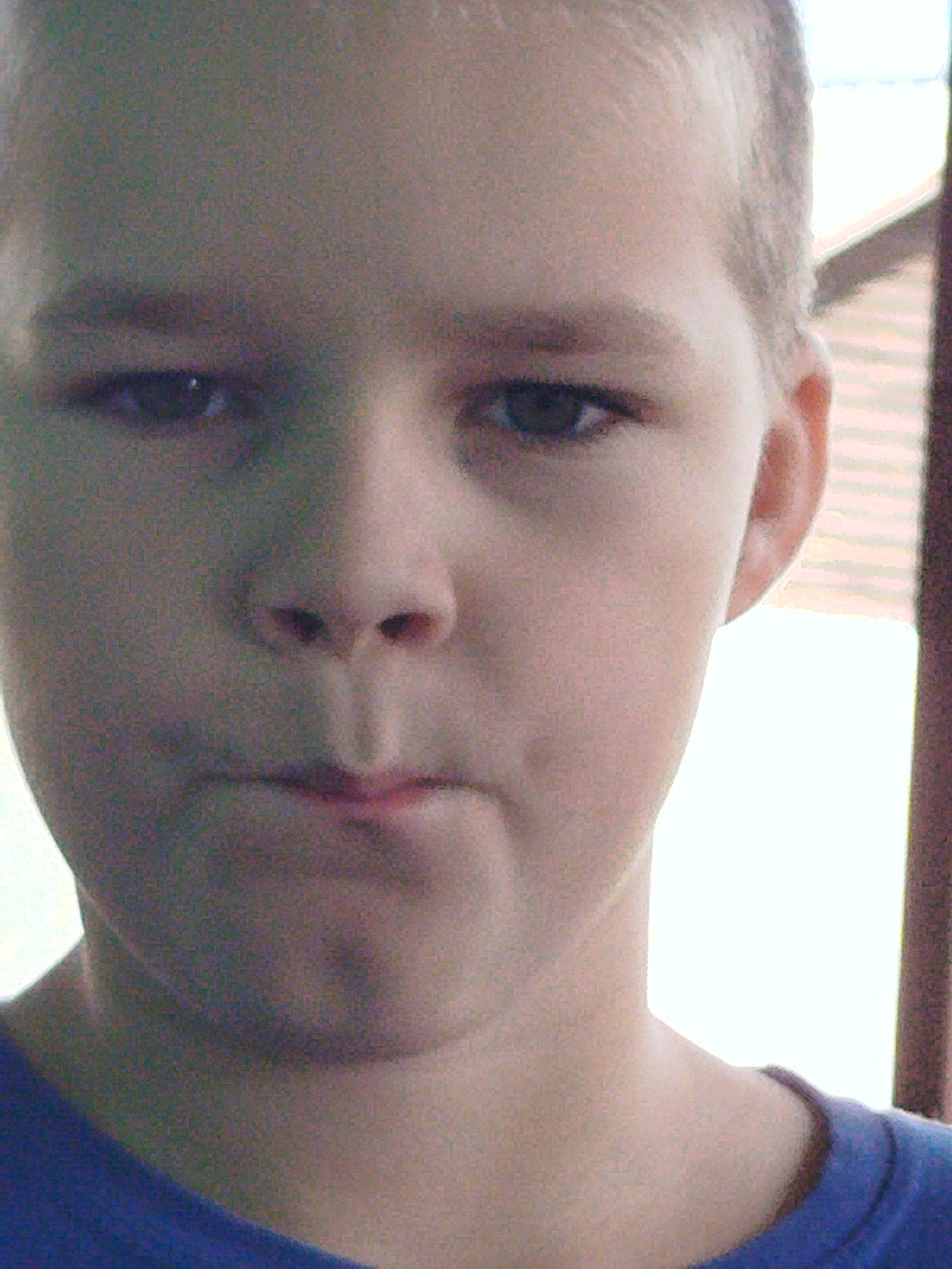 The image is a close-up, low-resolution portrait of a young person's face, likely a child, against a blurred background. The subject is facing the camera directly, with their head slightly tilted. The lighting is uneven, with the left side of the face being brighter than the right. The subject's skin tone appears fair. Their eyes are open and looking forward. The nose is prominent, and the lips are slightly parted. A hint of the neck and shoulder is visible, wearing a blue garment. The background is indistinct, featuring horizontal lines that could suggest blinds or slats, and a bright, out-of-focus area. The overall impression is of a casual, possibly candid, photograph taken indoors or in a shaded outdoor area. The location in Крывасельскі сельскі Савет, Belarus is not discernible from the visual information provided in the image.