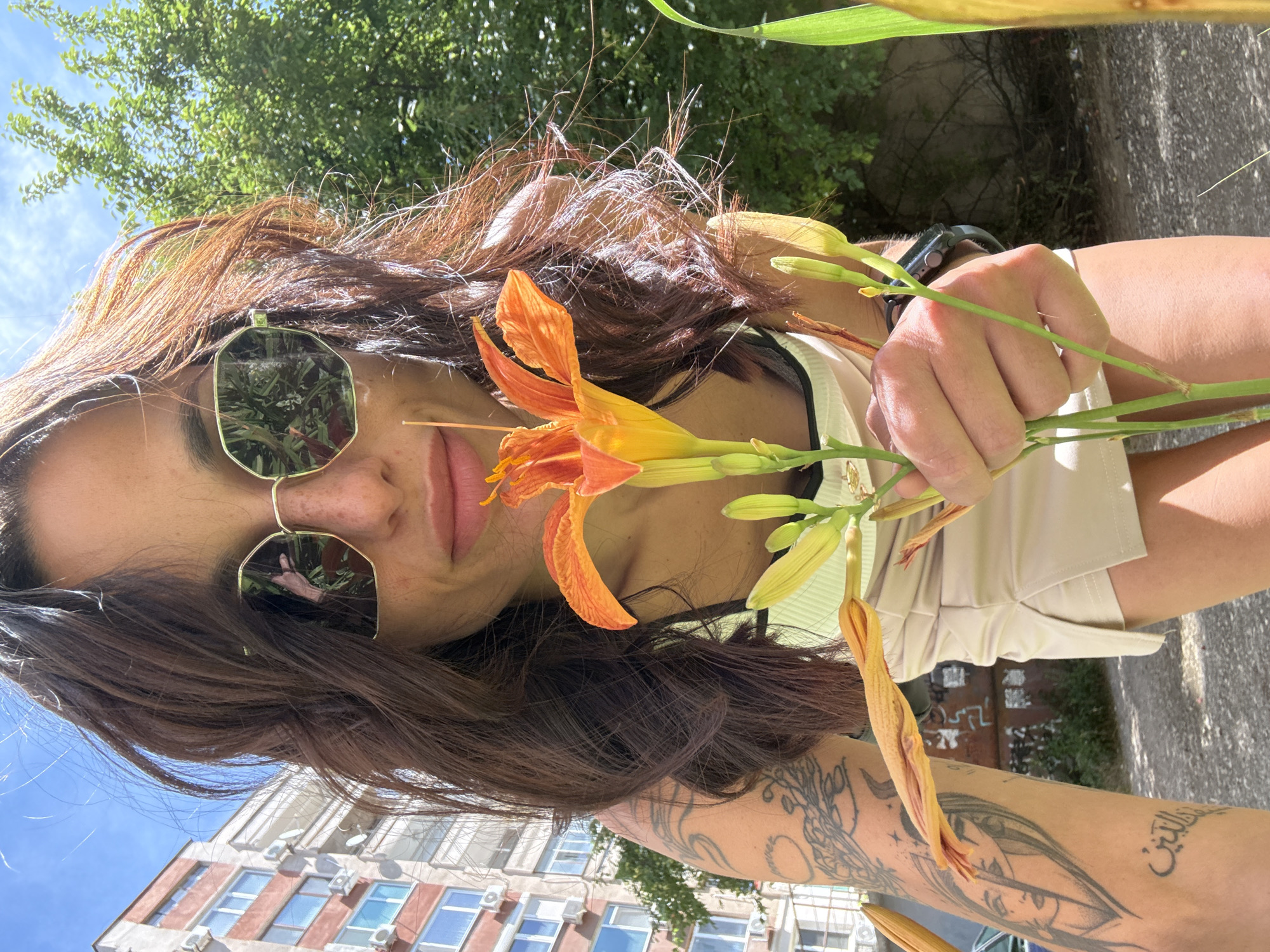 A young woman with brown hair and sunglasses is holding an orange lily in front of her face. She is wearing a light-colored tank top and a light-colored skirt. She has tattoos on her left arm. The background shows a building, trees, and a blue sky. The image was taken in the city of Pleven, Bulgaria, on a sunny day.