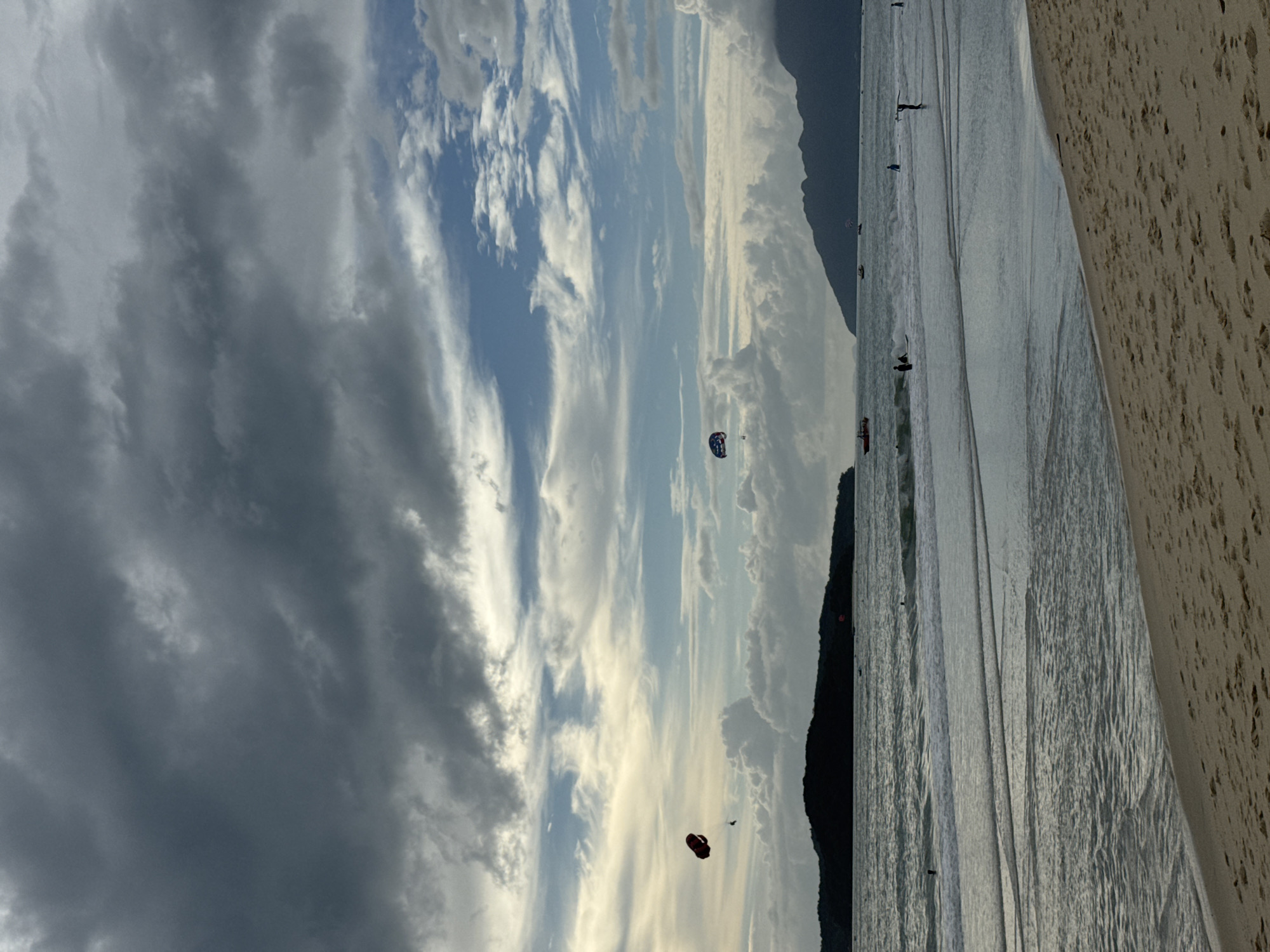 This image depicts a serene beach scene on Tengah Island, Malaysia, under a dramatic sky filled with a mix of dark and light clouds. The ocean is calm with gentle waves lapping onto a sandy shore. In the distance, silhouetted islands create a picturesque backdrop. Two parasails are visible in the sky, one red and one with a blue and white pattern, indicating recreational activity. Several small boats are scattered across the water, and a few individuals can be seen in the ocean, some possibly swimming or engaging in water sports. The foreground is dominated by a sandy beach, showing footprints and texture. The overall atmosphere is one of tranquility with a touch of adventurous spirit due to the parasailing.
