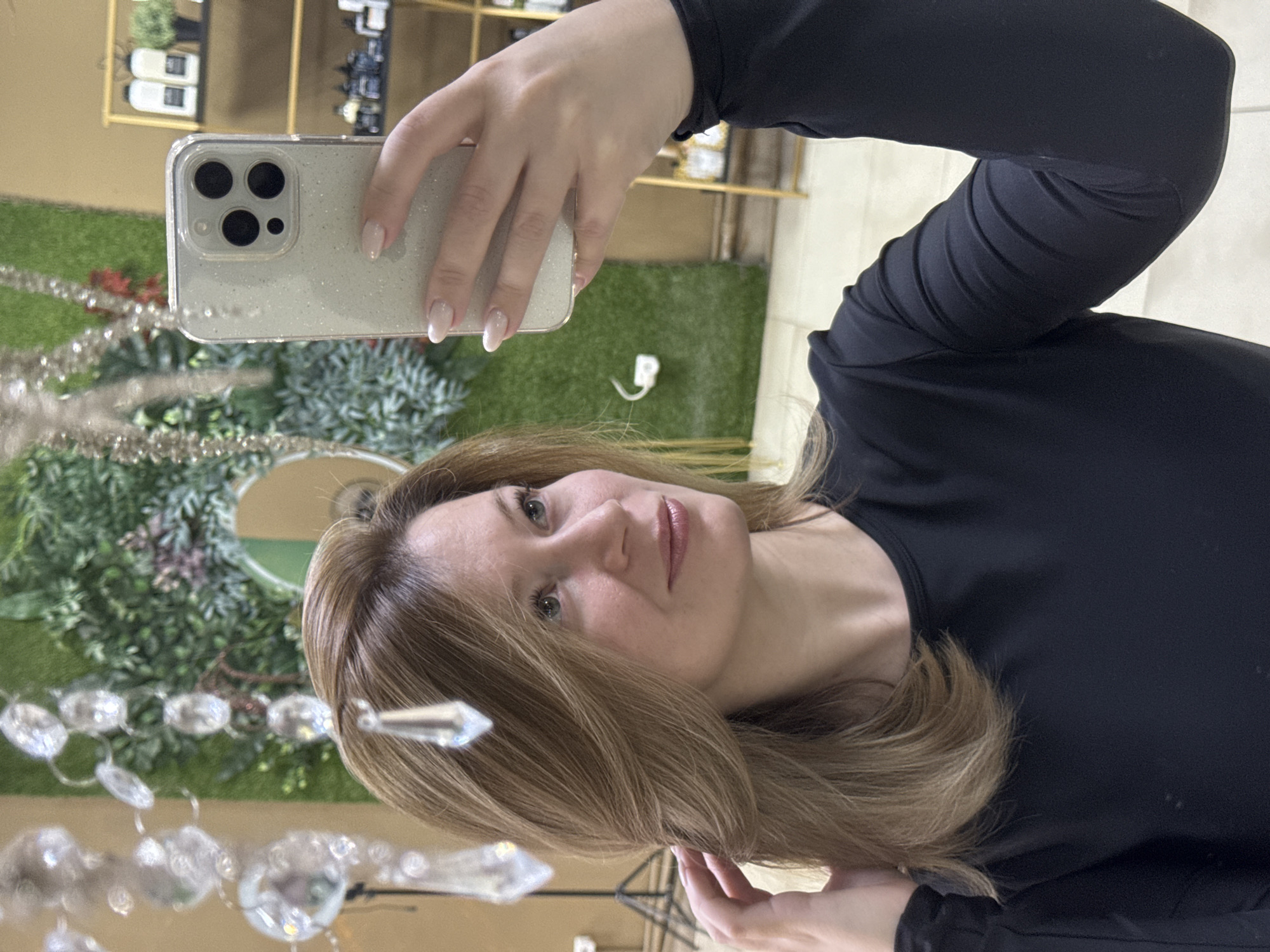 A woman is taking a selfie indoors. She is wearing a black long-sleeved shirt and holding a smartphone with a glittery case. The background includes a wall with green decor, a shelf with objects, and hanging crystals. The location may be a store or salon. The specific location is Chaikovsky, Russia.