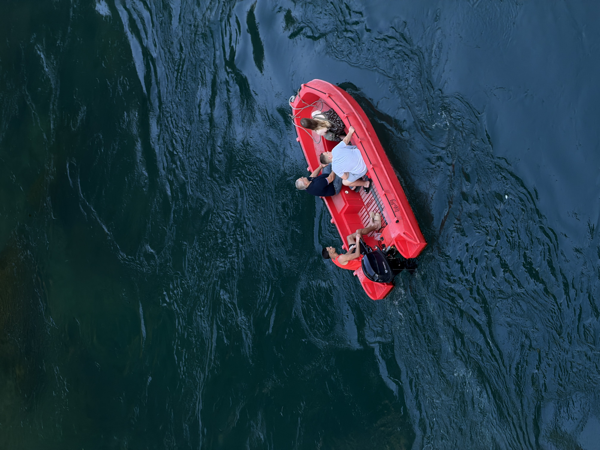 Four individuals are aboard a bright red inflatable boat with an outboard motor. The boat is positioned on dark, choppy water. In the stern, a person wearing a red tank top and black shorts operates the boat. Ahead of them, three passengers are seated. The individual in the front passenger seat wears a light blue t-shirt and grey shorts. The person next to them is wearing a dark blue t-shirt and jeans. The fourth person, a woman, is seated further back and wears a patterned dress. The boat appears to be moving, as indicated by the ripples in the water. This scene is located on the Neretva River in Mostar, Bosnia and Herzegovina, likely near the iconic Old Bridge, given the common tourist activities in that area. The water's deep, dark color suggests depth and possibly strong currents. The overall impression is one of leisure activity on a river.
