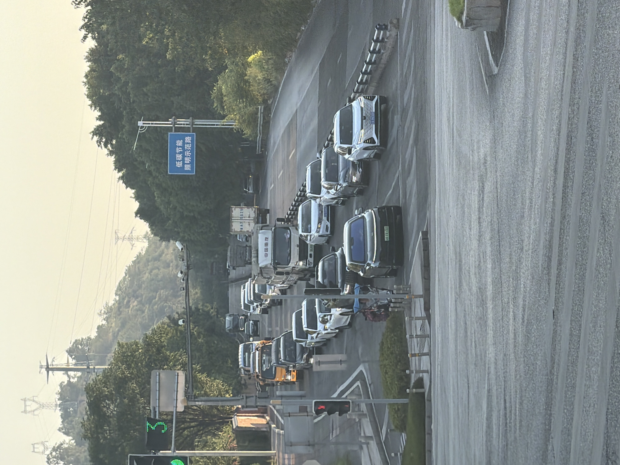 The image shows a street scene in Jiangjin, China, on a clear day with overcast skies. There is heavy traffic, with cars and trucks lined up on a multi-lane road. A traffic light is visible, showing a red signal. Power lines and towers are present in the background, along with trees and hills. A blue sign with Chinese characters is also visible on a pole. The road surface appears wet, and there are some reflections visible. A crosswalk is marked on the road.