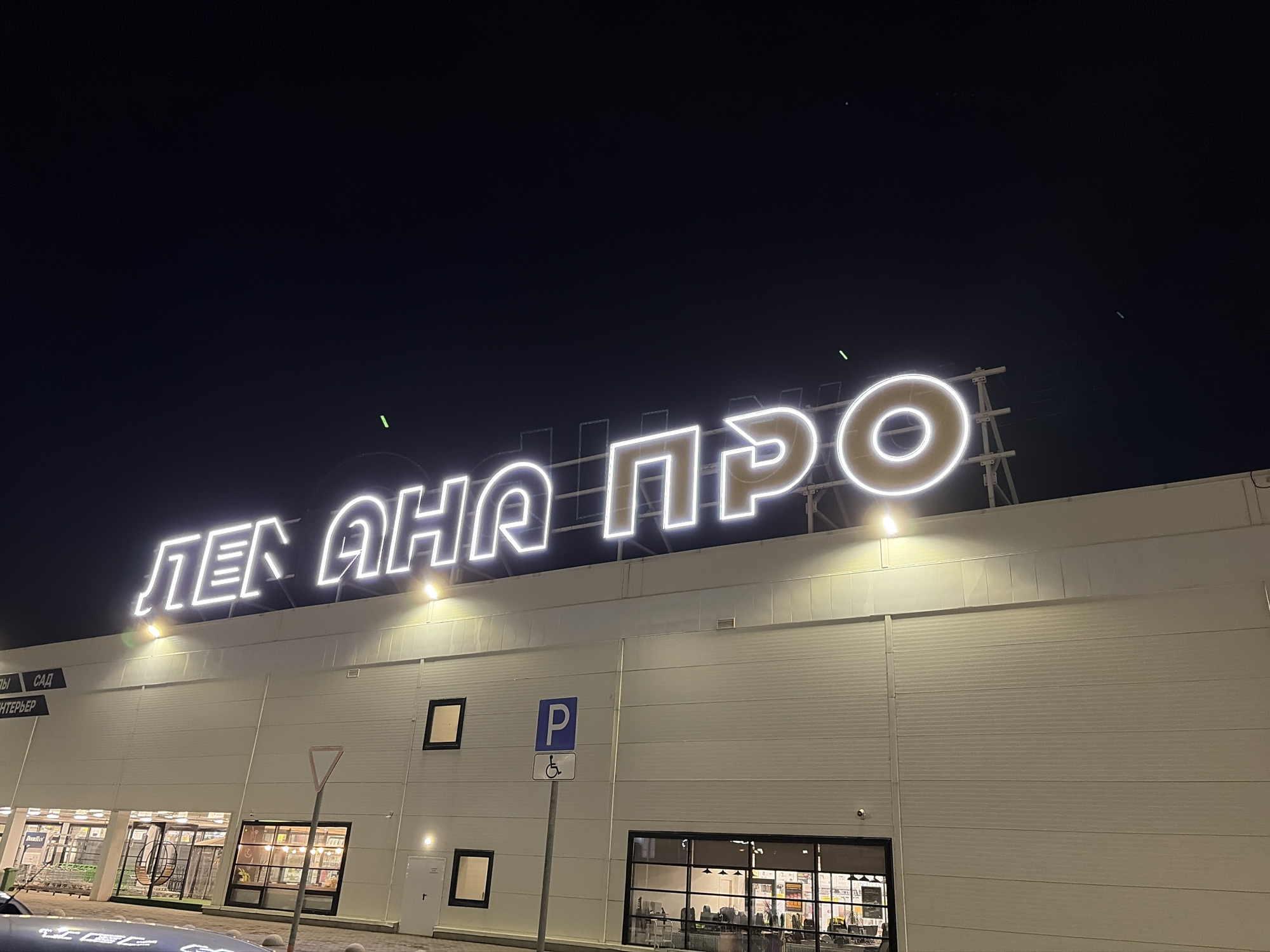 The image shows a building at night with a large, illuminated sign on top that reads "ЛЕН АНА ПРО". Based on the sign, the building appears to be a branch of Leroy Merlin, a home improvement and gardening multinational retailer. The building is white with several windows and a parking sign with a disabled symbol. The location is Izhevsk, Russia, as indicated by the Cyrillic alphabet used in the sign. There are no people visible in the image.