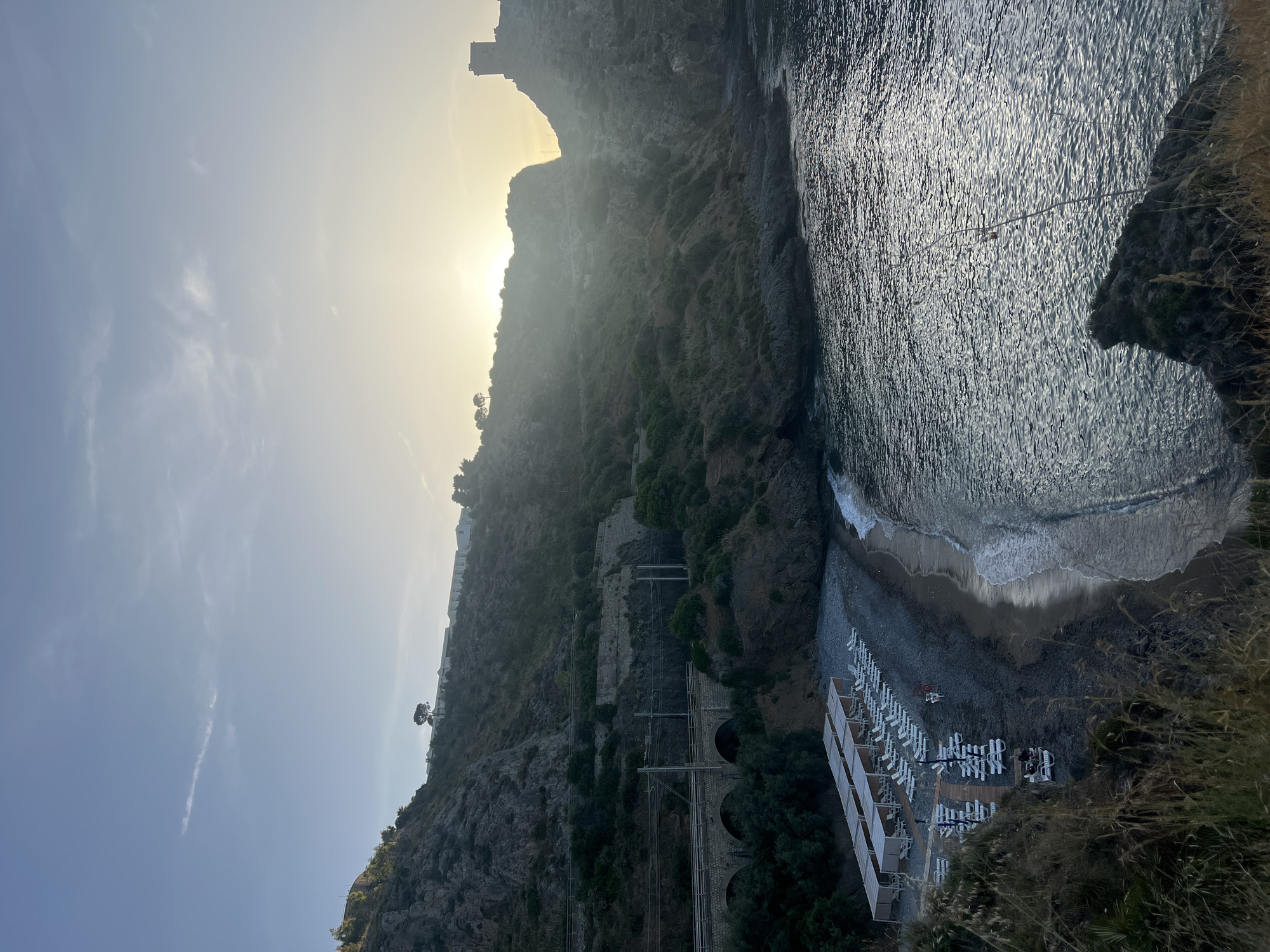 The image depicts a coastal scene in Sperone, Italy, under low sunlight. A pebble beach, situated within a cove, is flanked by steep, vegetated cliffs. On the left cliff, a double-track railway line, equipped with overhead electrical wires, traverses a three-arch stone bridge. A series of white, rectilinear buildings are visible along the distant ridge of this left cliff.
The right cliff rises steeply, culminating in a prominent dark, square stone tower at its summit, silhouetted against the bright, low sun. The sky is light blue with sparse, wispy clouds.
The beach itself is furnished with multiple rows of white lounge chairs. Several rectangular canopies provide shade over some of these chairs. Two individuals are seated in a lounge chair on a wooden platform in the lower left section of the beach. Two additional individuals are present further along the beach: one in a red top standing near the waterline, and another in a white top walking inland. The sea, to the right, displays a dark, shimmering surface with ripples reflecting the sky and sun, and gentle waves break along the shore. Dry grasses and foliage occupy the immediate lower left foreground.