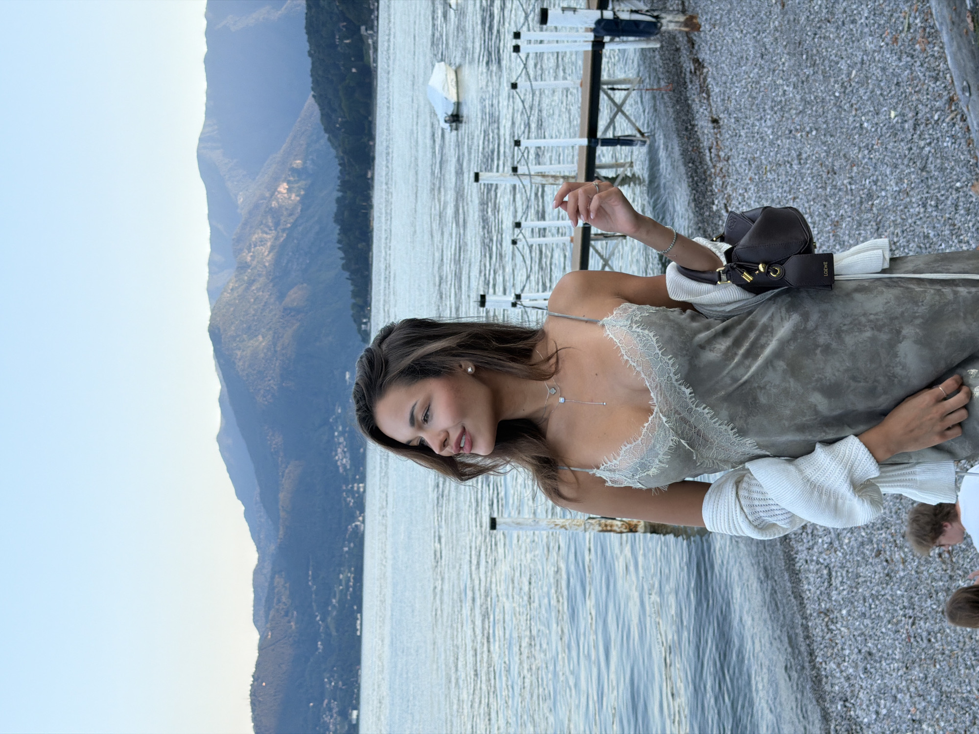 A young woman stands on a pebble beach near a lake in Nobiallo, Italy. She is wearing a light grey slip dress with lace detailing, a white cardigan draped over her shoulders, and a dark brown handbag. The lake is calm, with a few boats visible in the distance. Mountains form the backdrop, with a clear blue sky overhead. Two other individuals are partially visible in the lower left corner of the frame, seated on the beach.