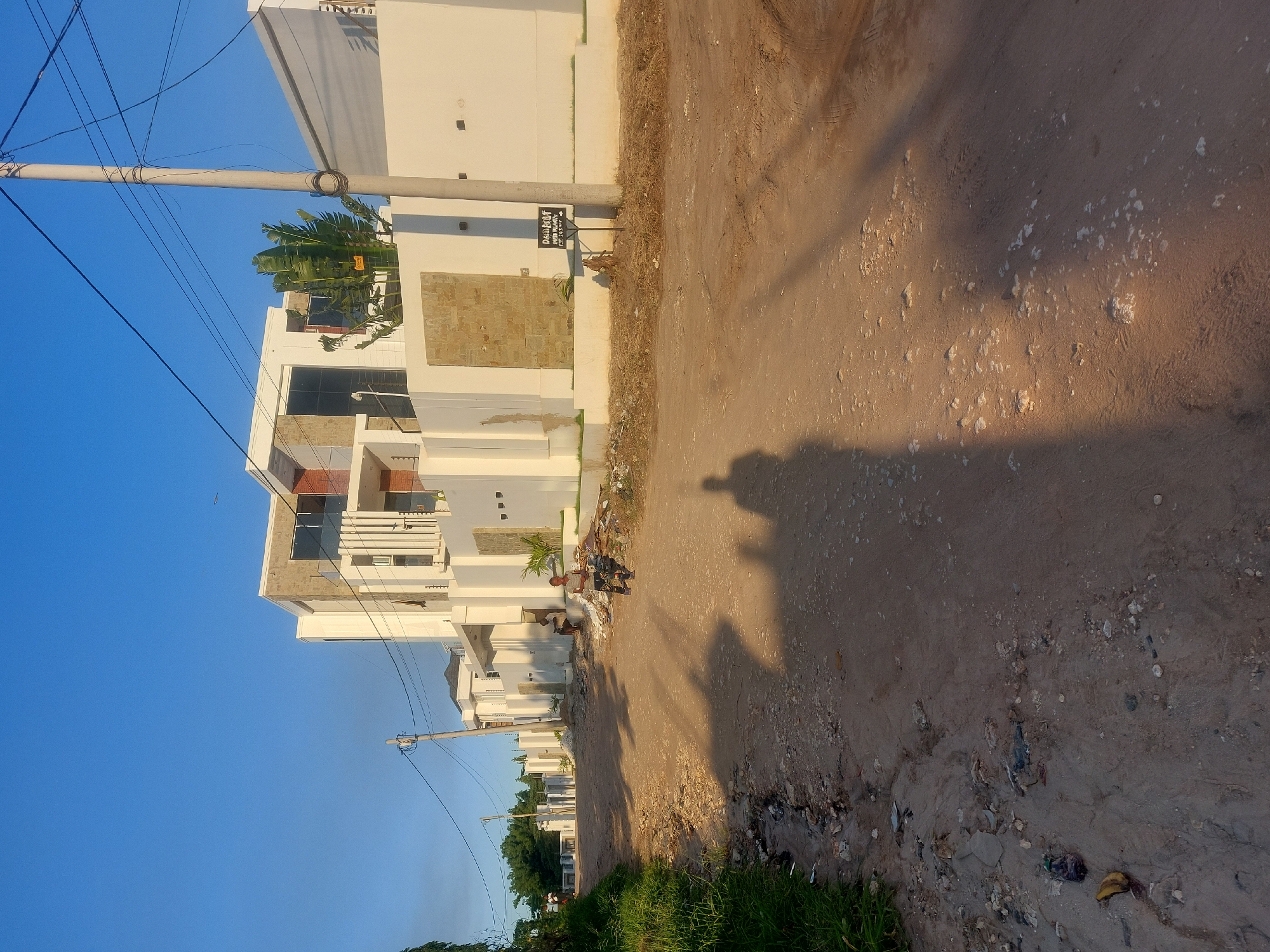 The scene shows a dirt road in Dar es-Salaam, Tanzania, lined with modern, multi-story buildings. A utility pole with multiple wires is visible against a clear blue sky. Debris and trash are scattered along the roadside. Two people are standing in front of one of the buildings. The shadow of the photographer is cast on the dirt road.