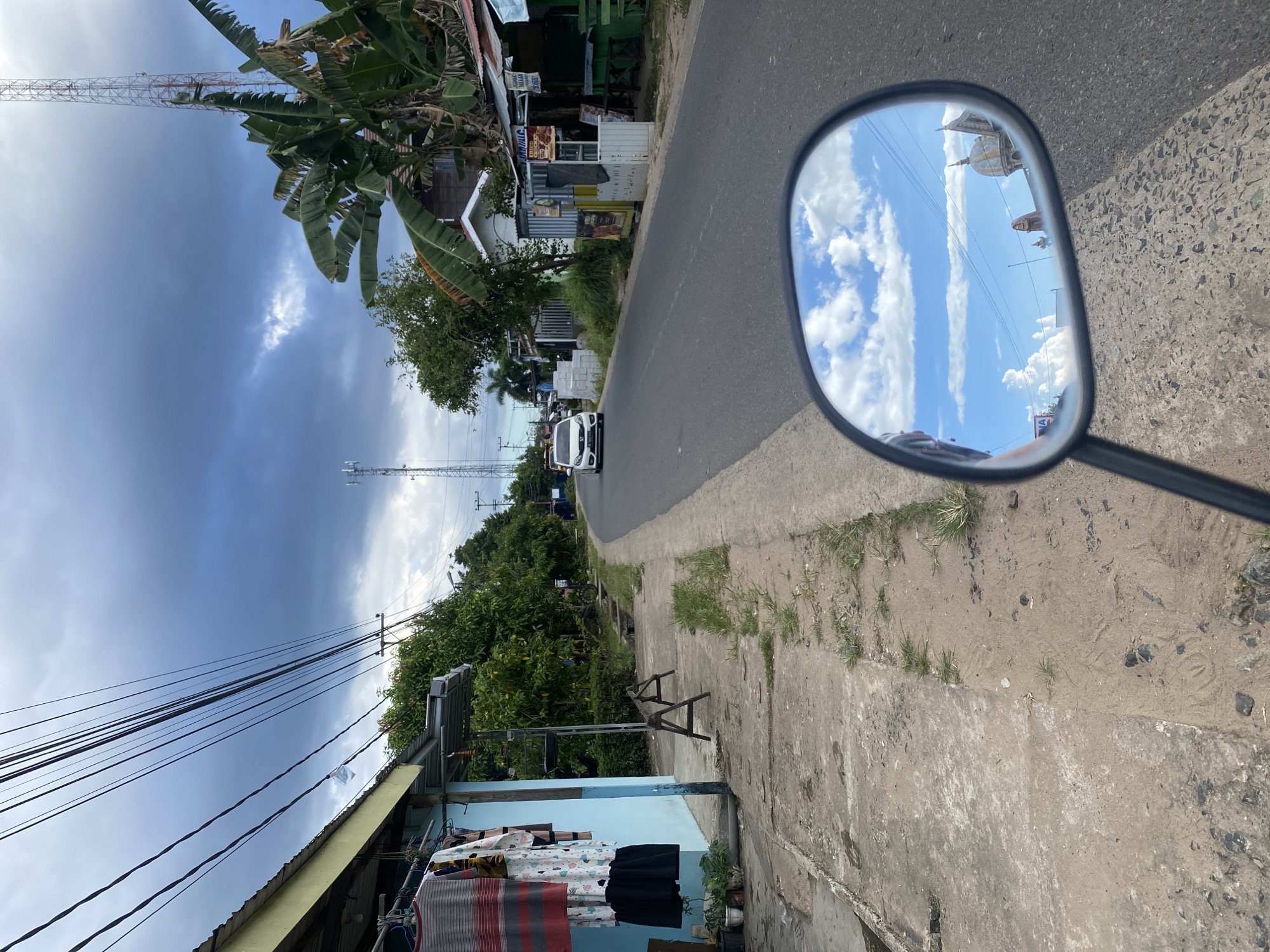 The image captures a street scene in Sungai Sipai, Indonesia, viewed from the perspective of someone riding a motorcycle. A light-colored car is driving on the asphalt road, which curves to the right. On the left side of the road, a light blue building with a yellow overhang is visible. Clothes are hanging on a line in front of the building. A sidewalk made of concrete and dirt runs alongside the building, with some patches of grass growing through it. Further down the road, trees line the sides, and various structures, including what appear to be small shops and houses, are present. A tall telecommunications tower is visible in the distance. The sky is cloudy with patches of blue sky. A motorcycle's side mirror in the foreground reflects the sky and some building structures.