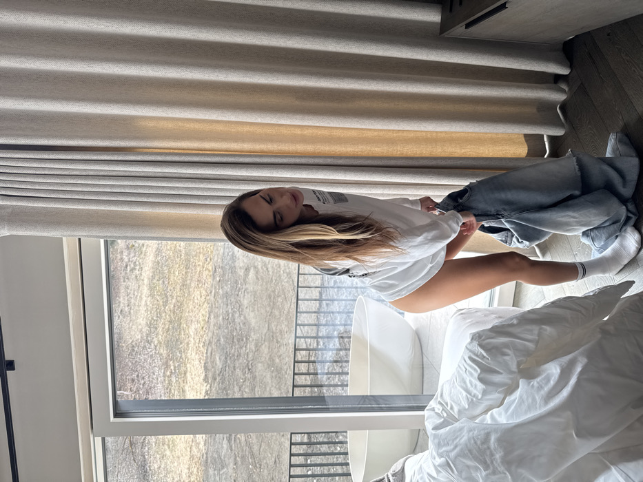 The image depicts a female subject indoors, within a modern accommodation in Schladming, Austria. She is captured in the process of handling light blue denim jeans, bending slightly forward with both hands on the garment. She wears an oversized white long-sleeved shirt, which covers her torso and upper thighs, and white socks with two thin black stripes at the ankle. Her long, light brown hair with blonde highlights is parted on the side and hangs over her shoulders.
In the background, a large floor-to-ceiling glass sliding door with a black frame offers a view to an outdoor patio or balcony. A white, freestanding oval bathtub is visible on this exterior space, positioned behind a dark metal railing. Beyond the patio, a muted, uncultivated landscape comprising grass and bare earth extends towards a distant, gently sloping hillside.
To the left of the subject, a portion of a bed with white, rumpled bedding is visible. To her right, light beige pleated curtains are partially drawn across the window, and a section of a dark wooden cabinet with a horizontal black handle is present. The room features dark wooden floorboards and a white ceiling equipped with a black track light fixture.