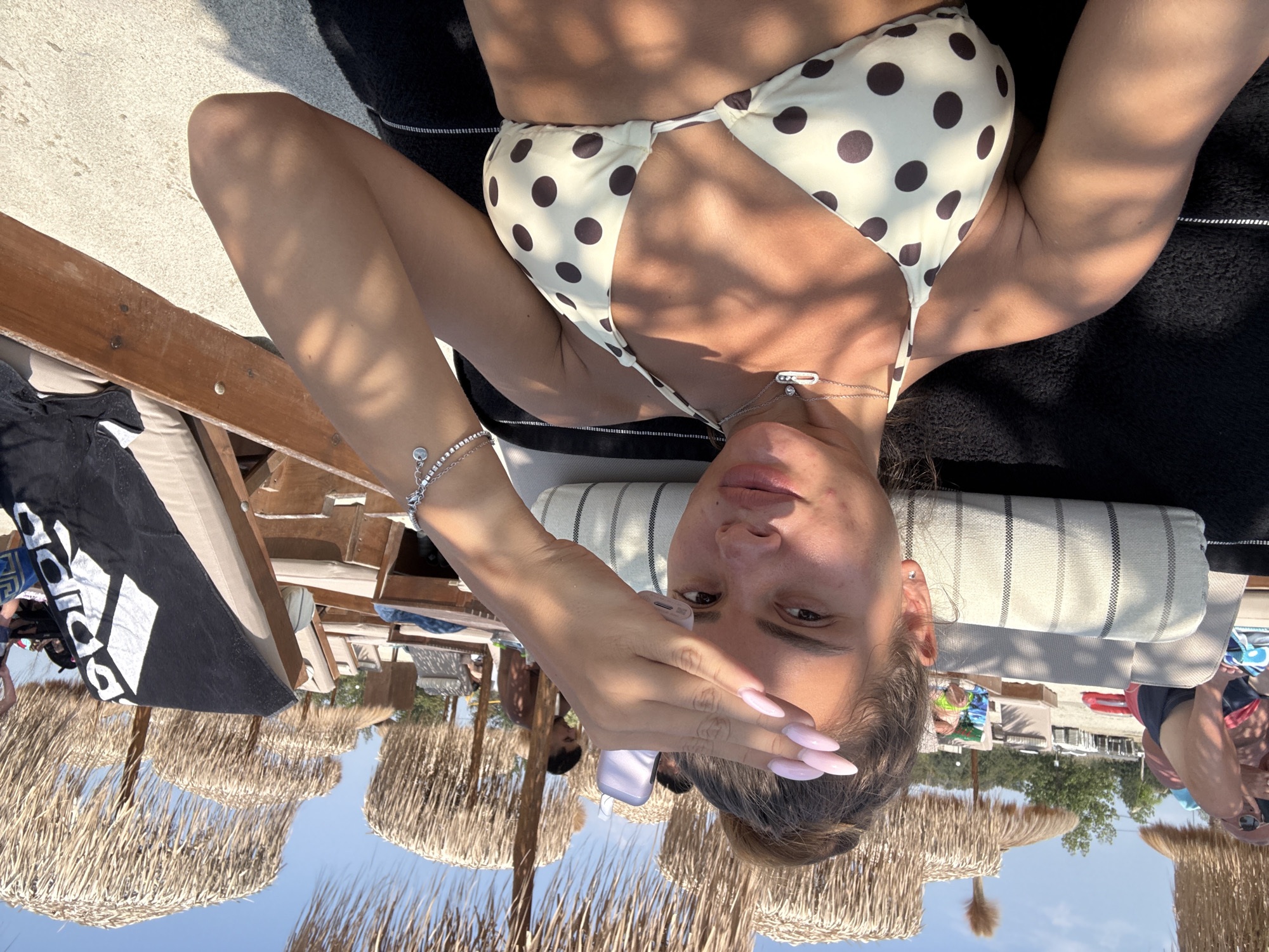 A young woman is reclining on a beach chair under a thatched umbrella, holding a small electronic device to shield her face from the sun. She is wearing a light-colored bikini with dark polka dots and a silver necklace with a pendant. Her right arm is raised, and her hand is positioned in front of her face. In the background, other beachgoers and umbrellas are visible. The setting appears to be a beach in the city of Katerini Municipality, Greece.