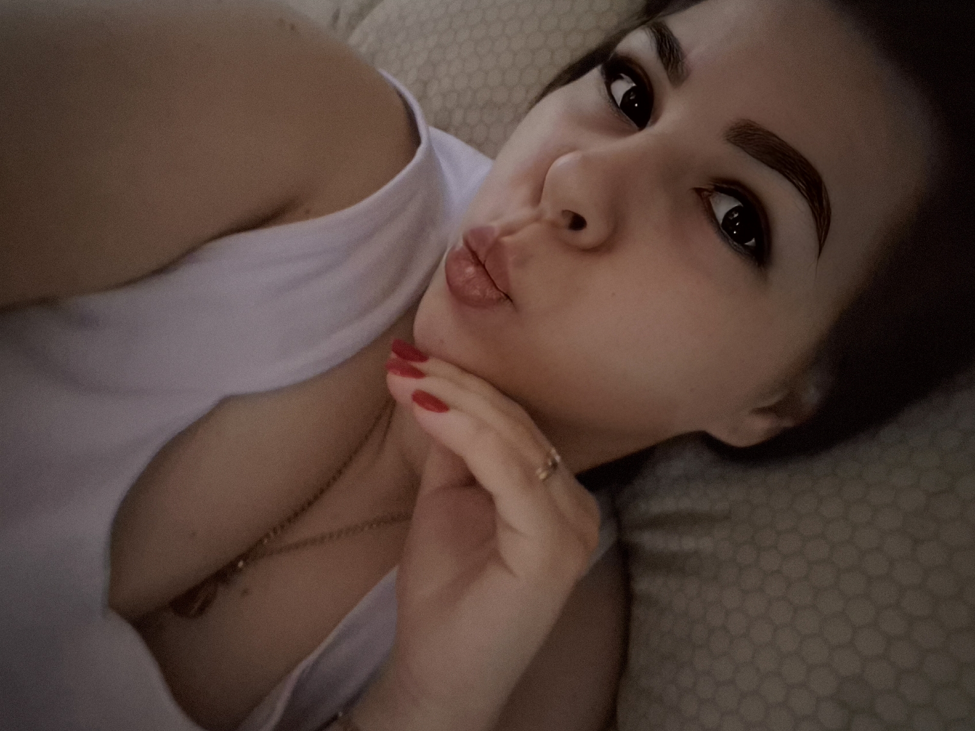 A woman is lying down, facing the camera with a pouting expression. She has dark hair and eyebrows and is wearing a white tank top. She rests her chin on her left hand, which has red nail polish and a ring on one finger. A gold necklace is visible around her neck. The background appears to be a textured cushion or bedding. The location is not discernible as Tobolsk, Russia.
